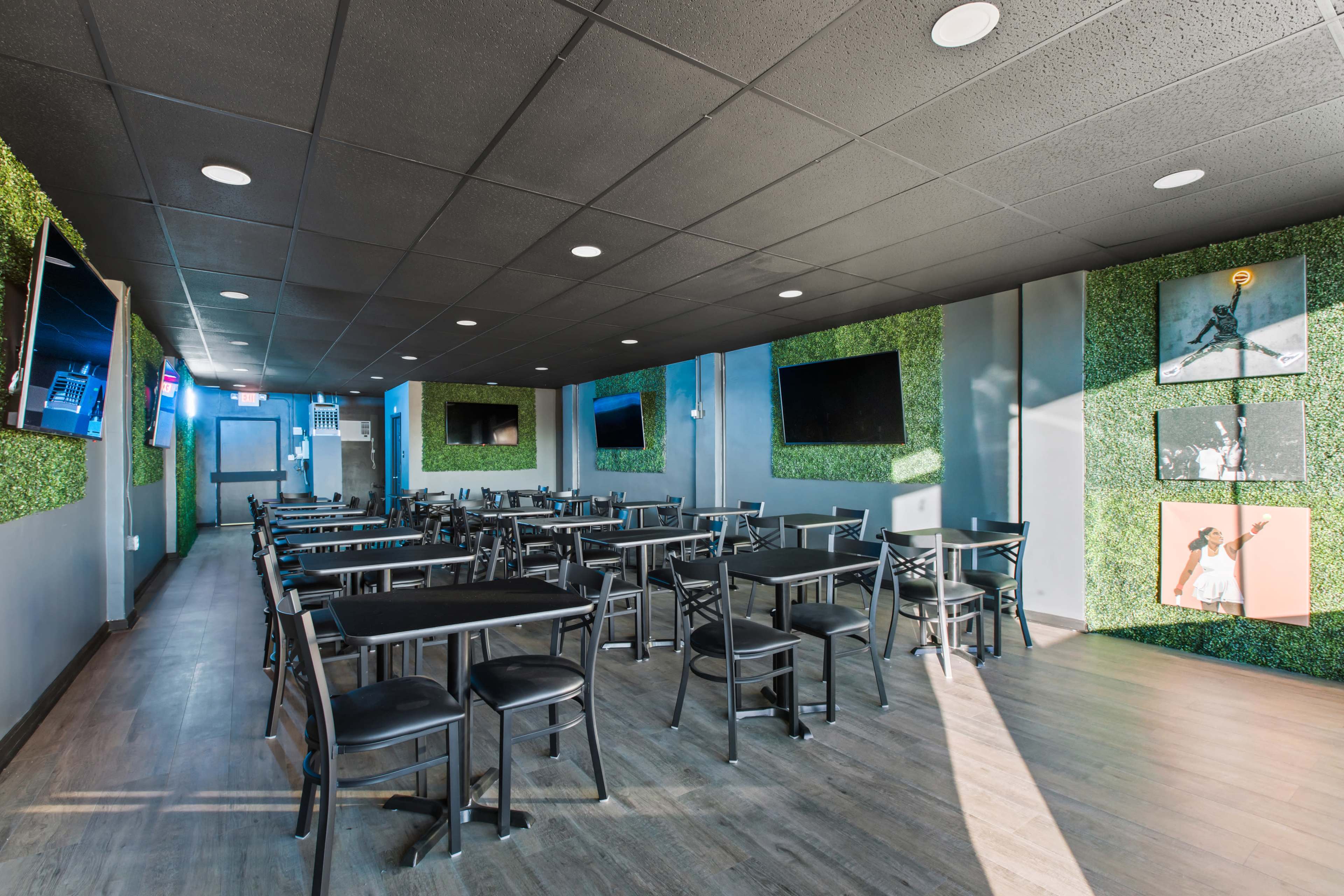 A spacious restaurant interior with black tables and chairs, green wall decor, and multiple televisions mounted on the walls.