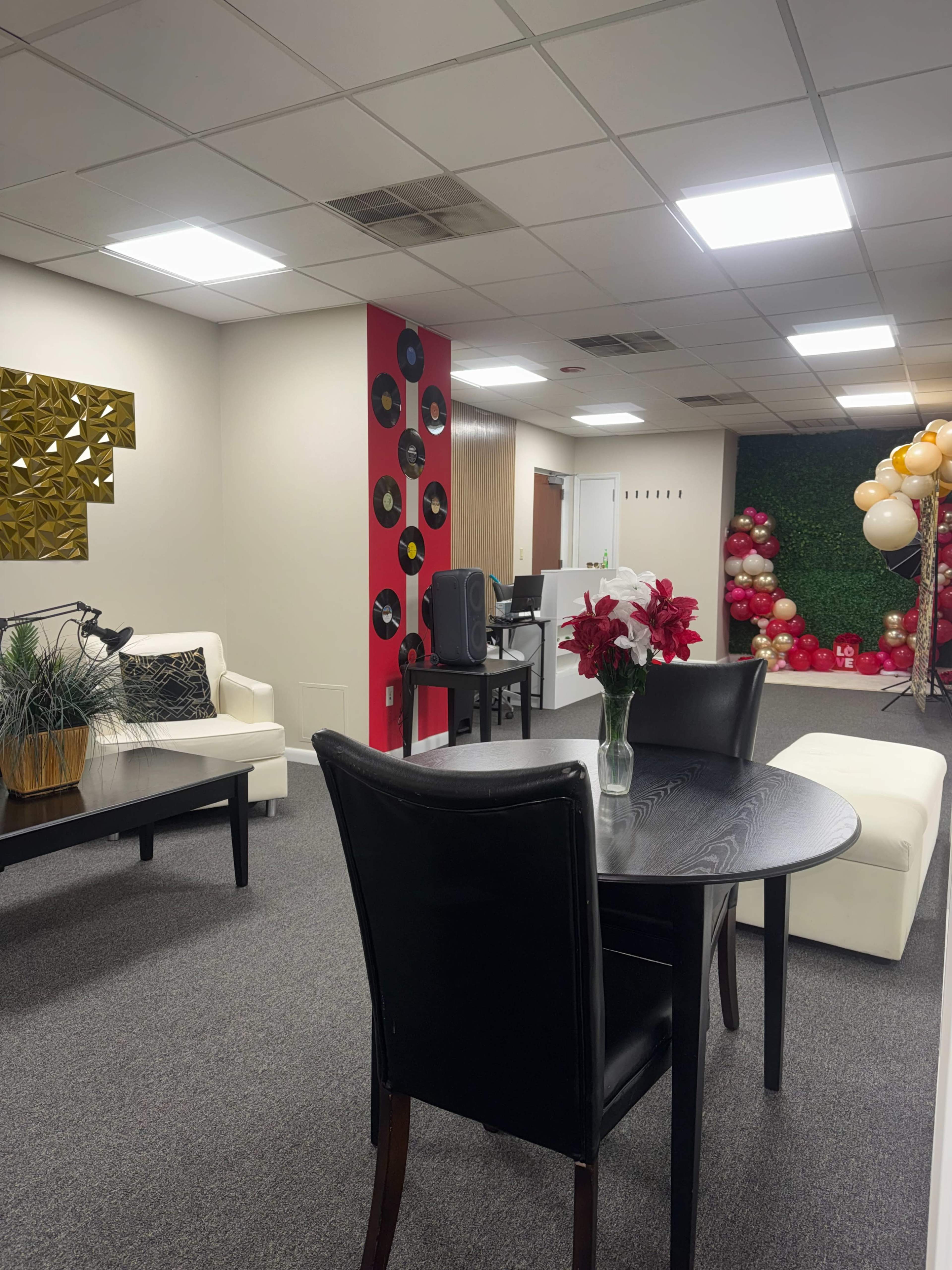 The image shows a modern office interior with a seating area featuring two white sofas, a round black dining table, and decorations including a wall with vinyl records and a corner adorned with balloons.