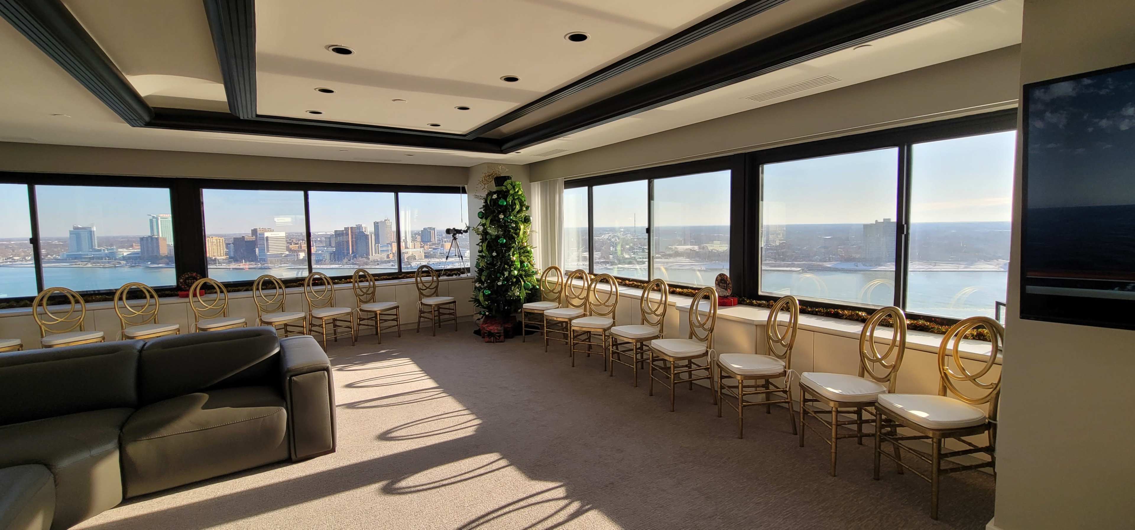 The image shows a spacious room with large windows offering a view of a city skyline and water, surrounded by several empty chairs arranged in a row.