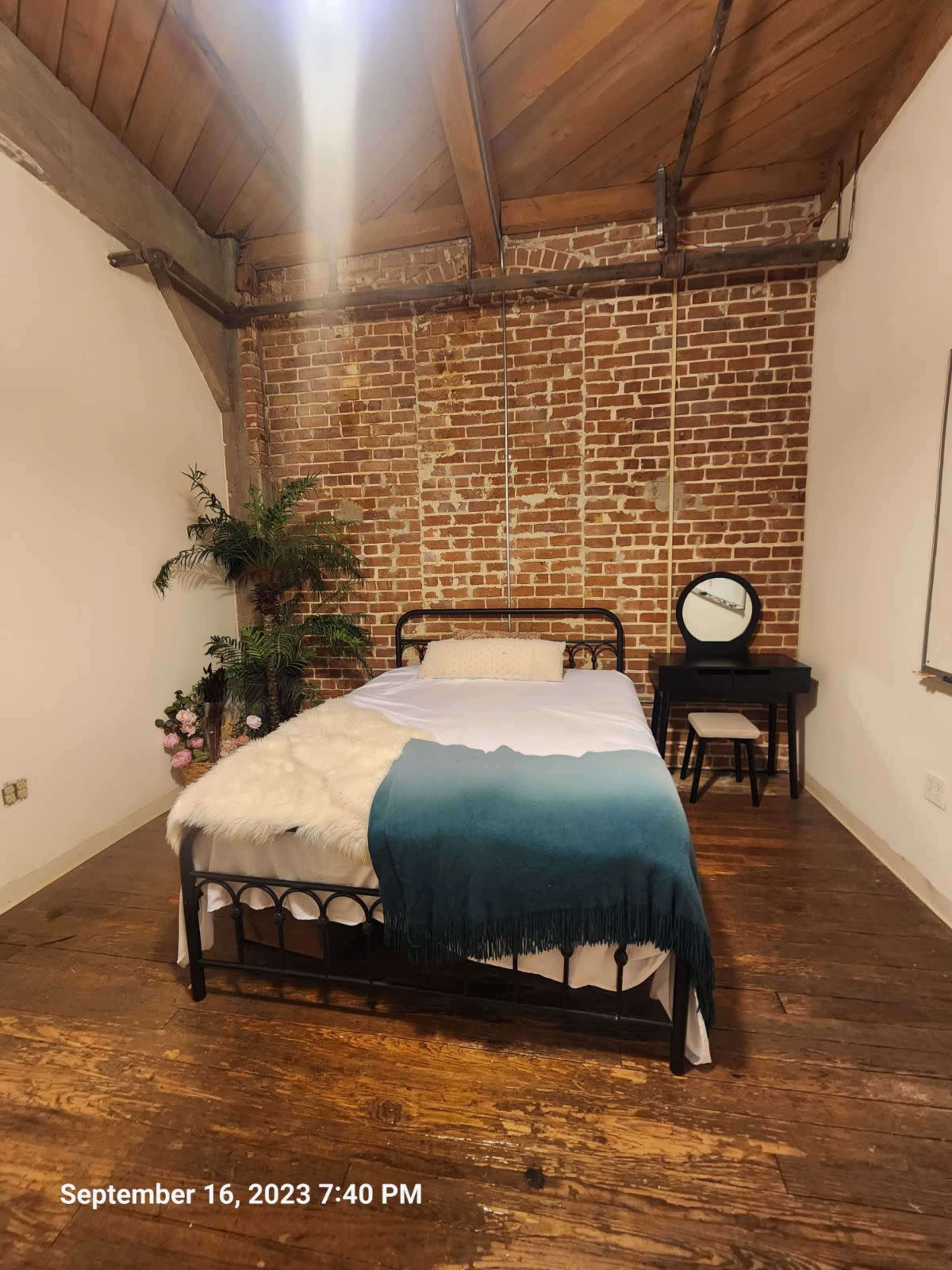 A bedroom with a black metal bed frame, a white sheet and a teal throw blanket, alongside a small desk with a mirror and potted plants against exposed brick walls.