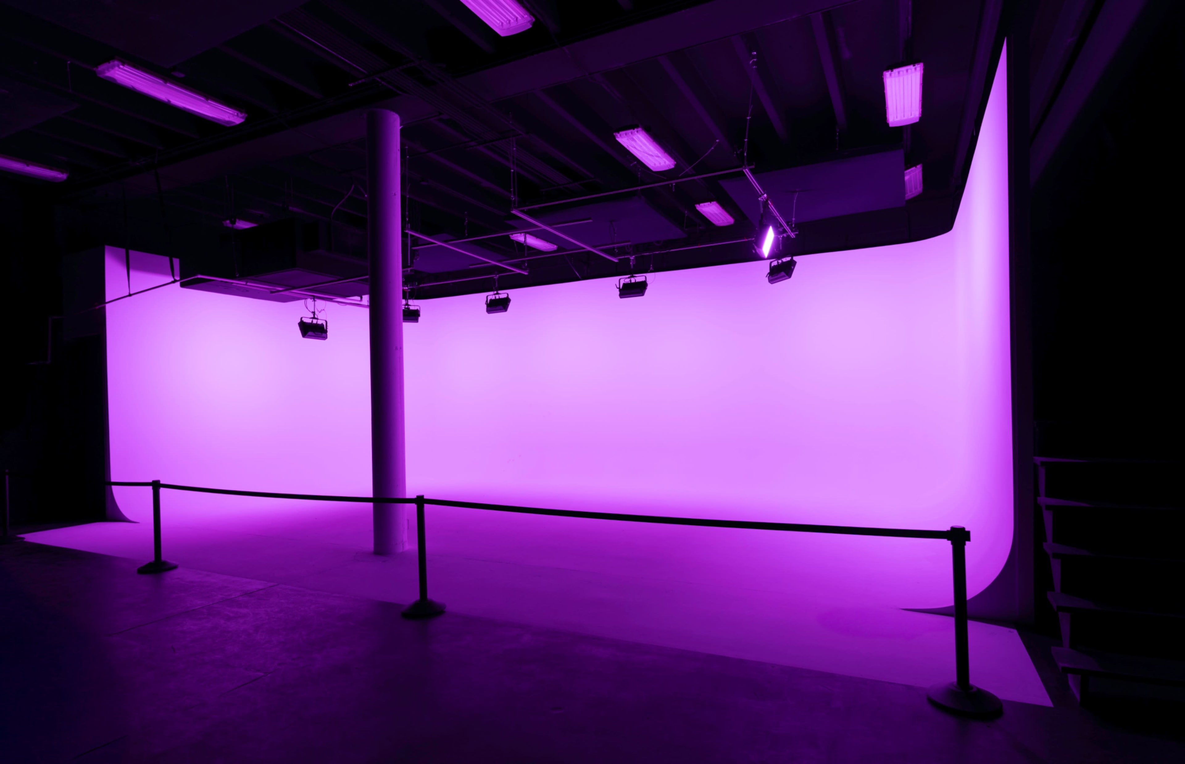 The image shows a minimalist studio space with a large curved backdrop illuminated in purple light and several spotlights positioned above.