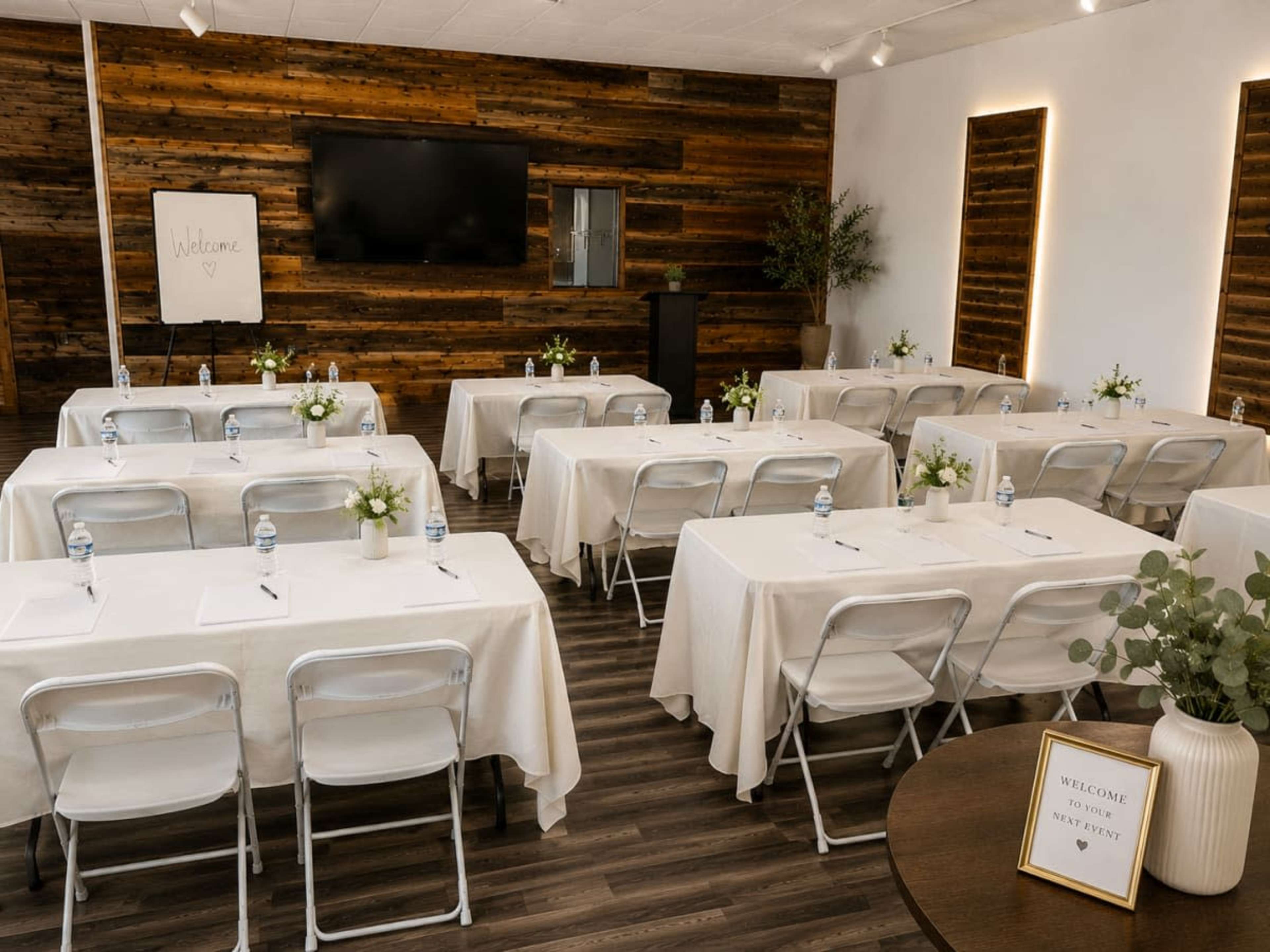 A classroom setting with several tables covered in white tablecloths, arranged in a U-shape, features water bottles and flower centerpieces, with a welcome sign and a television in the background.