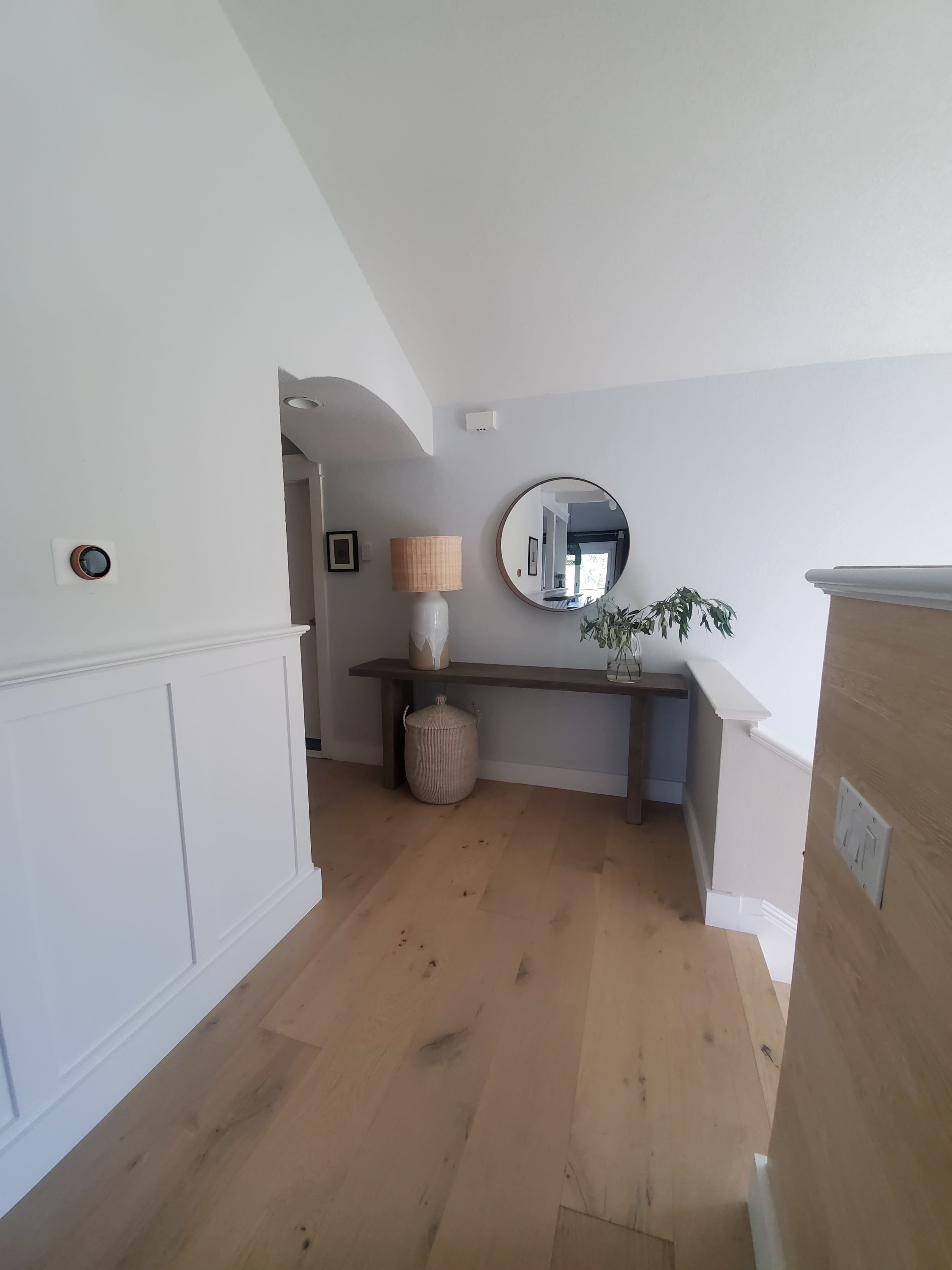 A hallway features a rounded mirror above a console table with a lamp and a small plant, set against white walls and a light wooden floor.