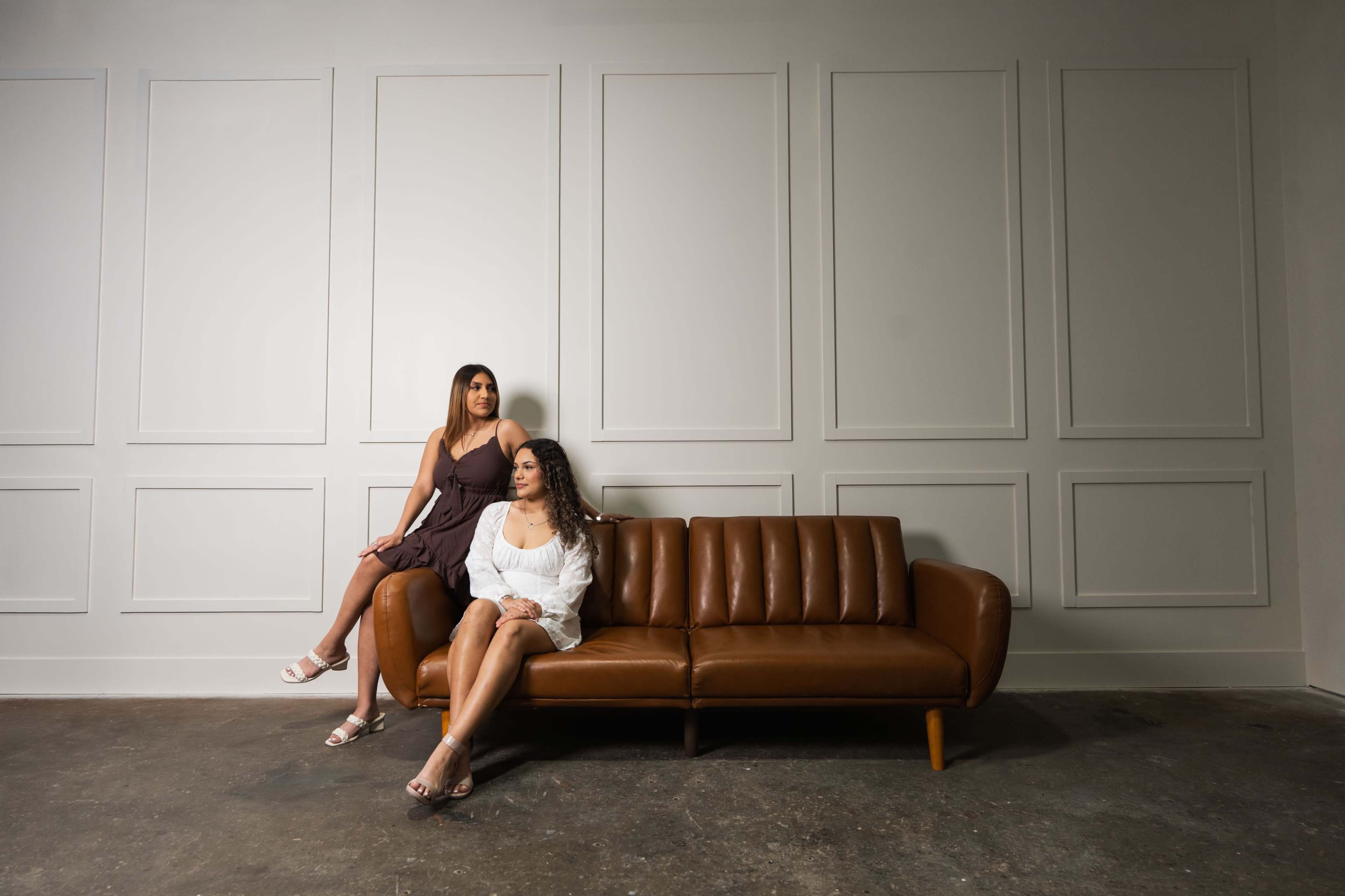 Two women sit on a brown leather sofa against a plain, light-colored wall in a minimalist room.