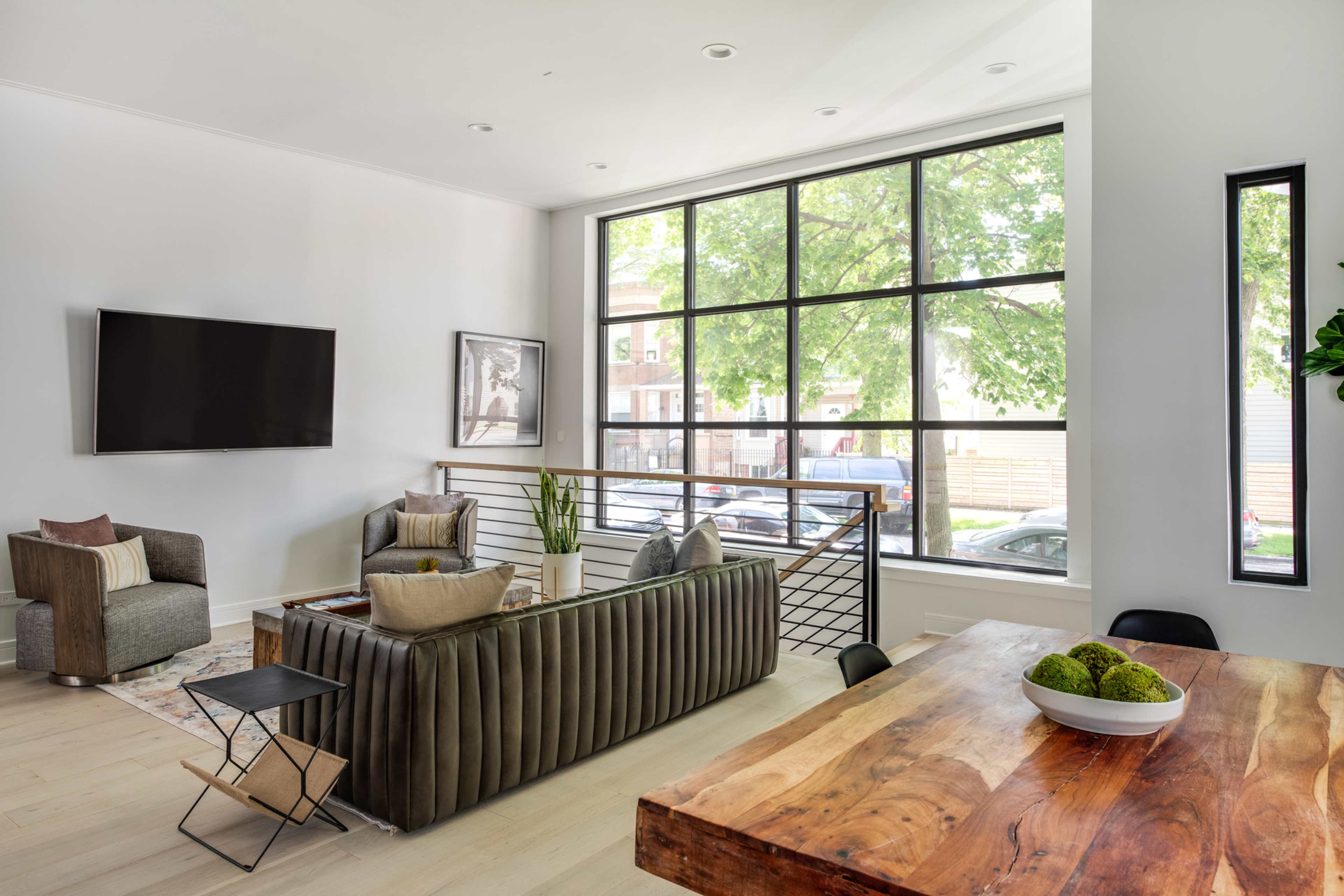 A modern living room features large windows, a gray sectional sofa, a wooden dining table, and minimalistic decor.