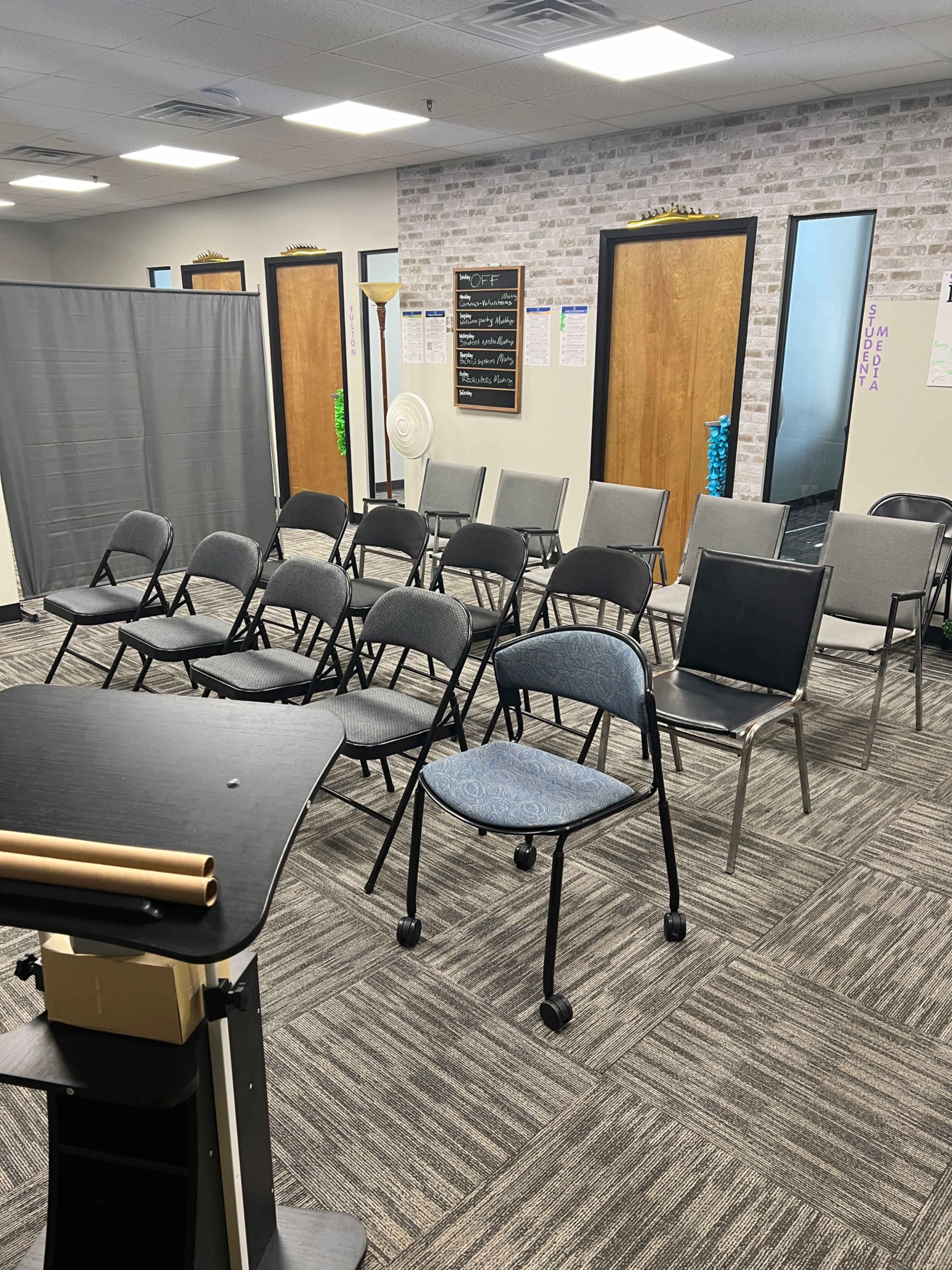 A meeting room with rows of folding chairs and a podium, featuring gray carpeting and a brick-patterned wall.