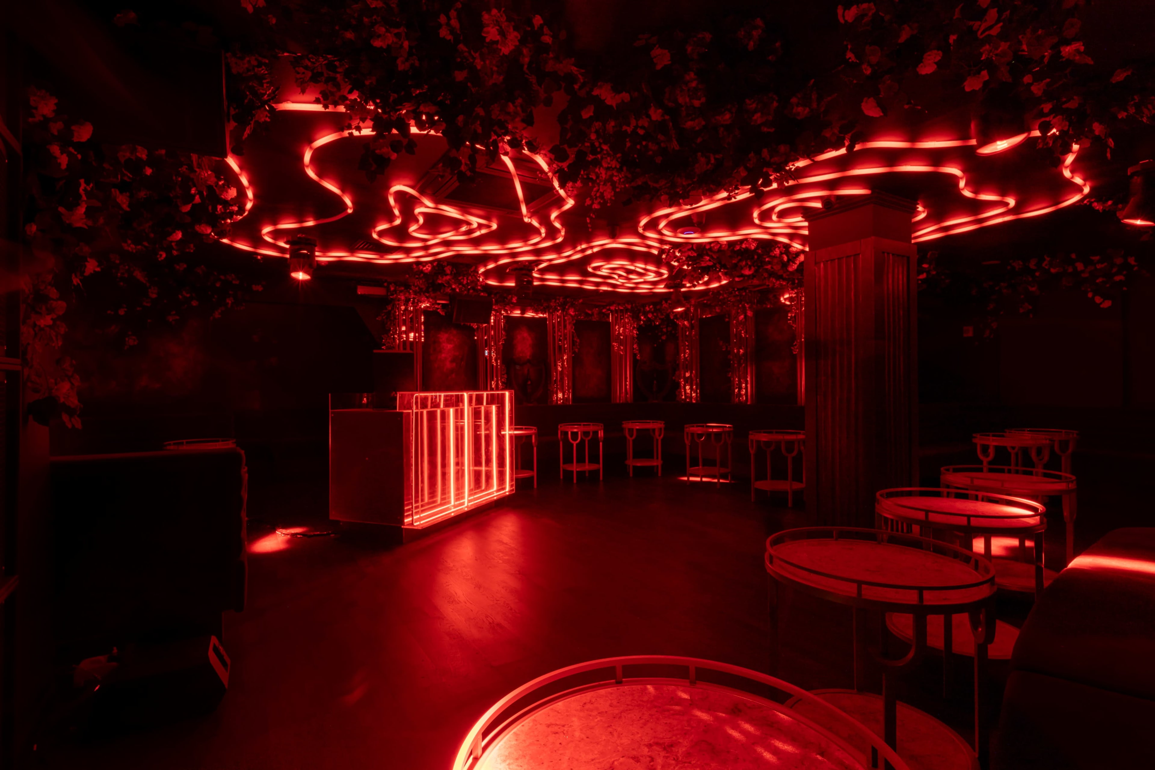 The interior of a dimly lit bar features red neon lighting, dark furnishings, and circular tables arranged around a central counter.