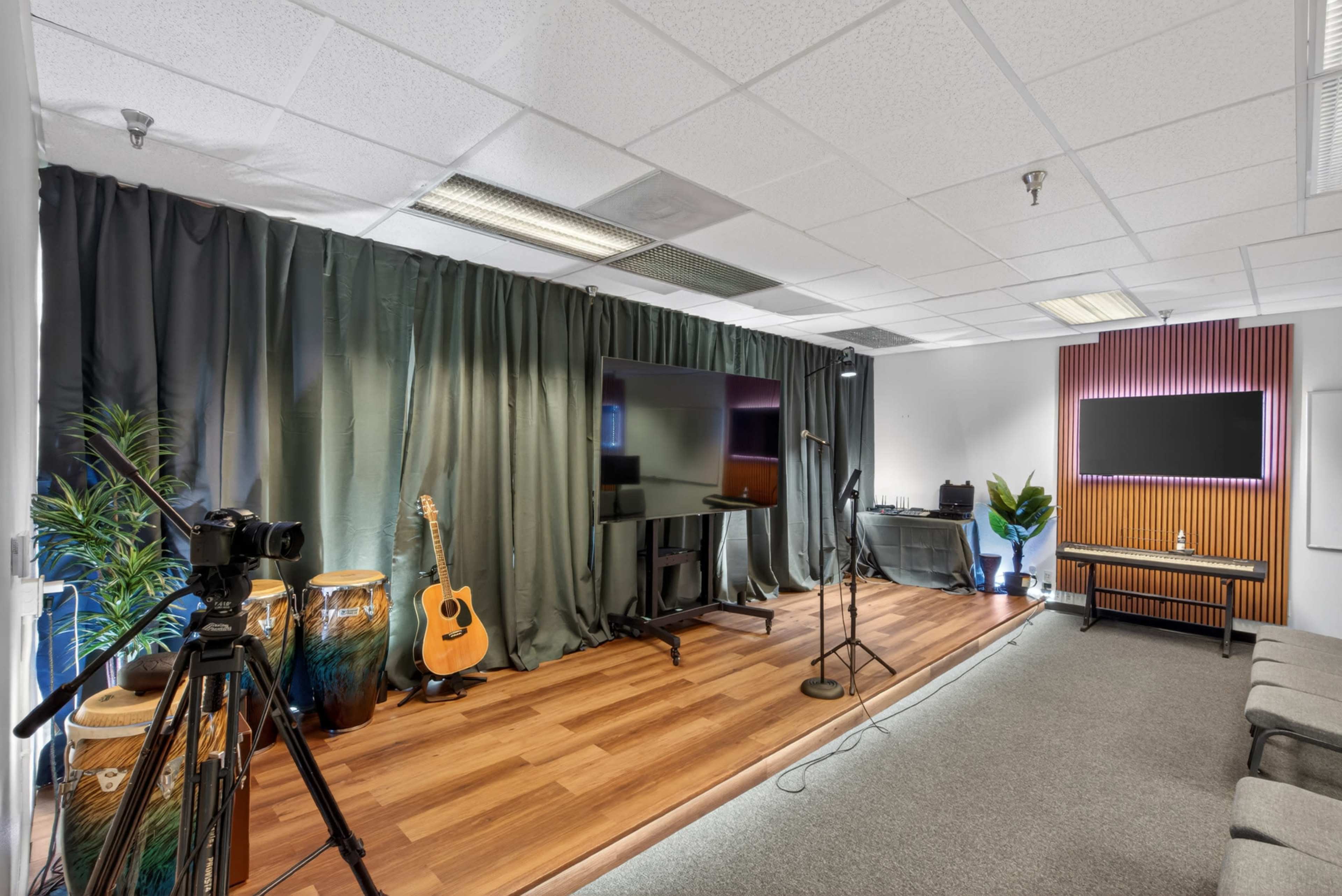 The image shows a modern studio setup featuring a microphone stand, a camera on a tripod, various percussion instruments, and two large screens against a curtain backdrop.