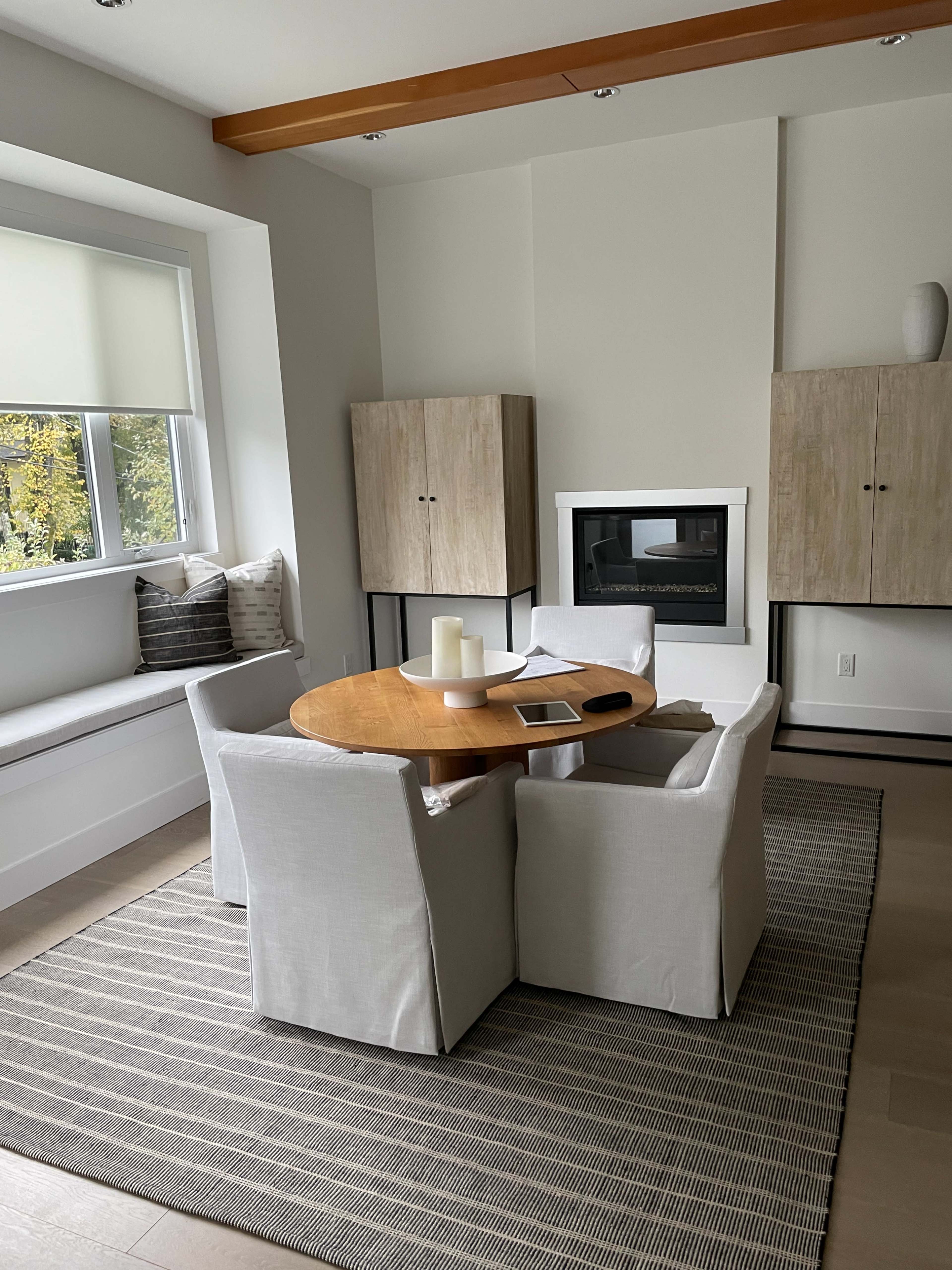 A round wooden table is set in a minimalist room, surrounded by four white upholstered chairs, with a window nook and a fireplace in the background.