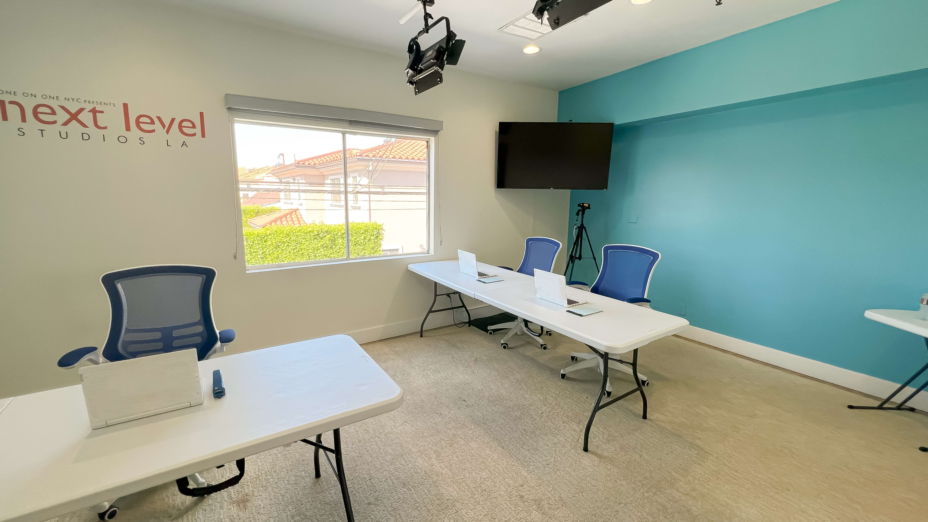 The image shows a small studio room with three tables arranged for meetings or presentations, each equipped with a laptop and a blue chair, and a large screen mounted on the wall.