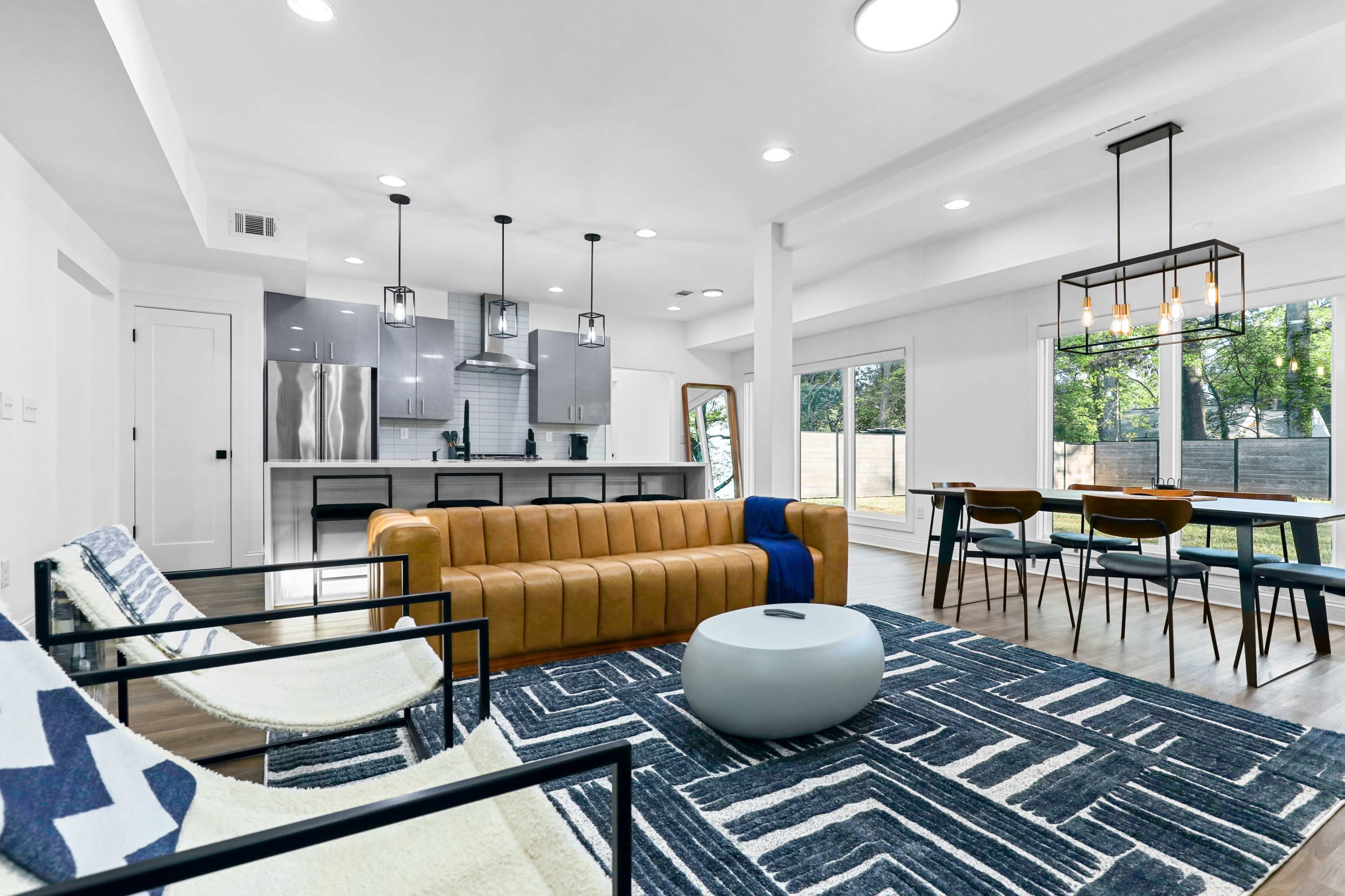 A modern living space with a brown leather couch, black chairs, a patterned rug, and a dining area with a wooden table and chairs, all set in front of large windows overlooking a yard.