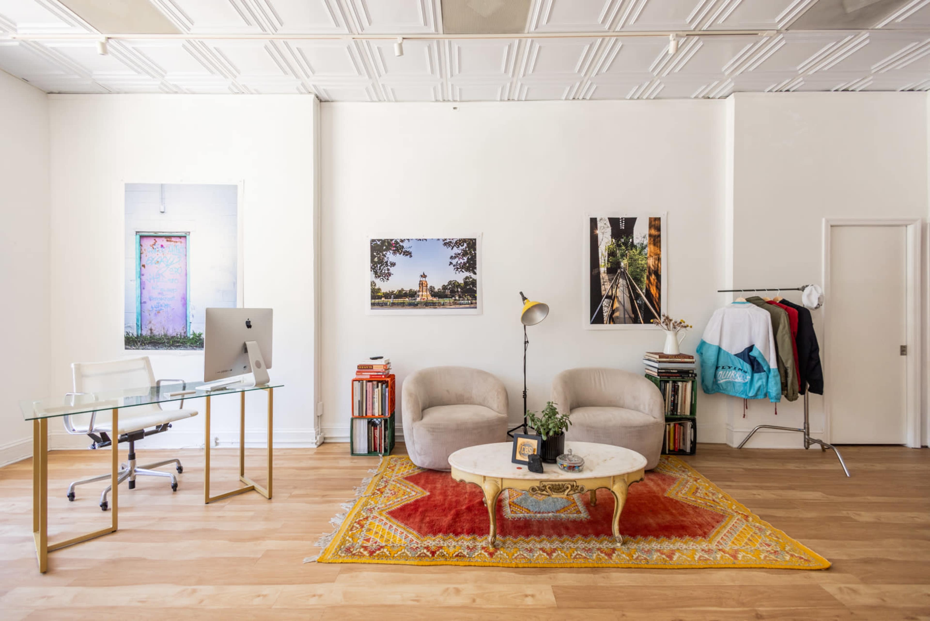 The image shows a modern office space featuring two beige armchairs, a glass desk with an iMac, a decorative rug, and framed photographs on the walls.