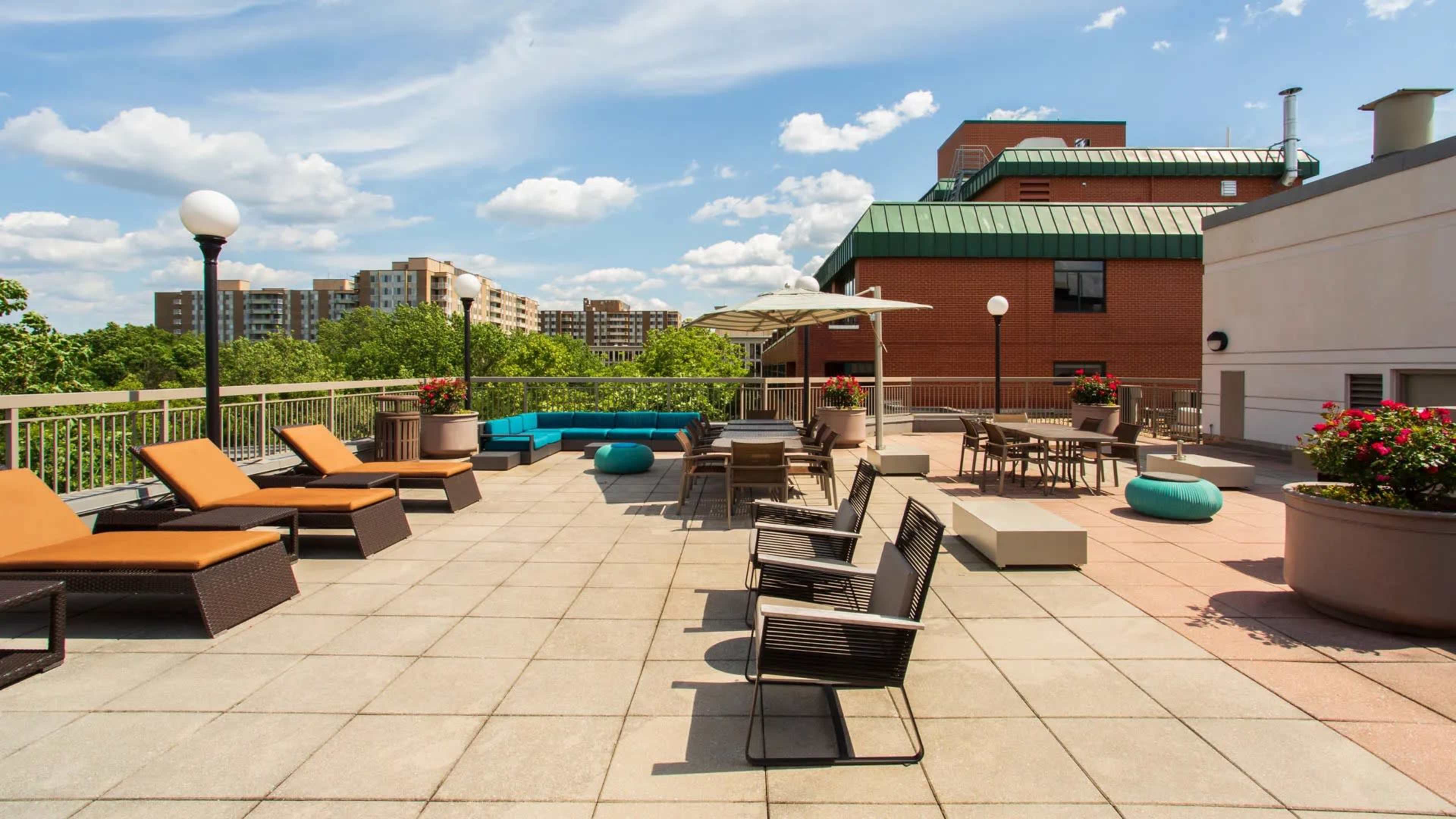 A spacious rooftop terrace with lounge chairs, tables, and an umbrella, overlooking a cityscape with greenery in the background.