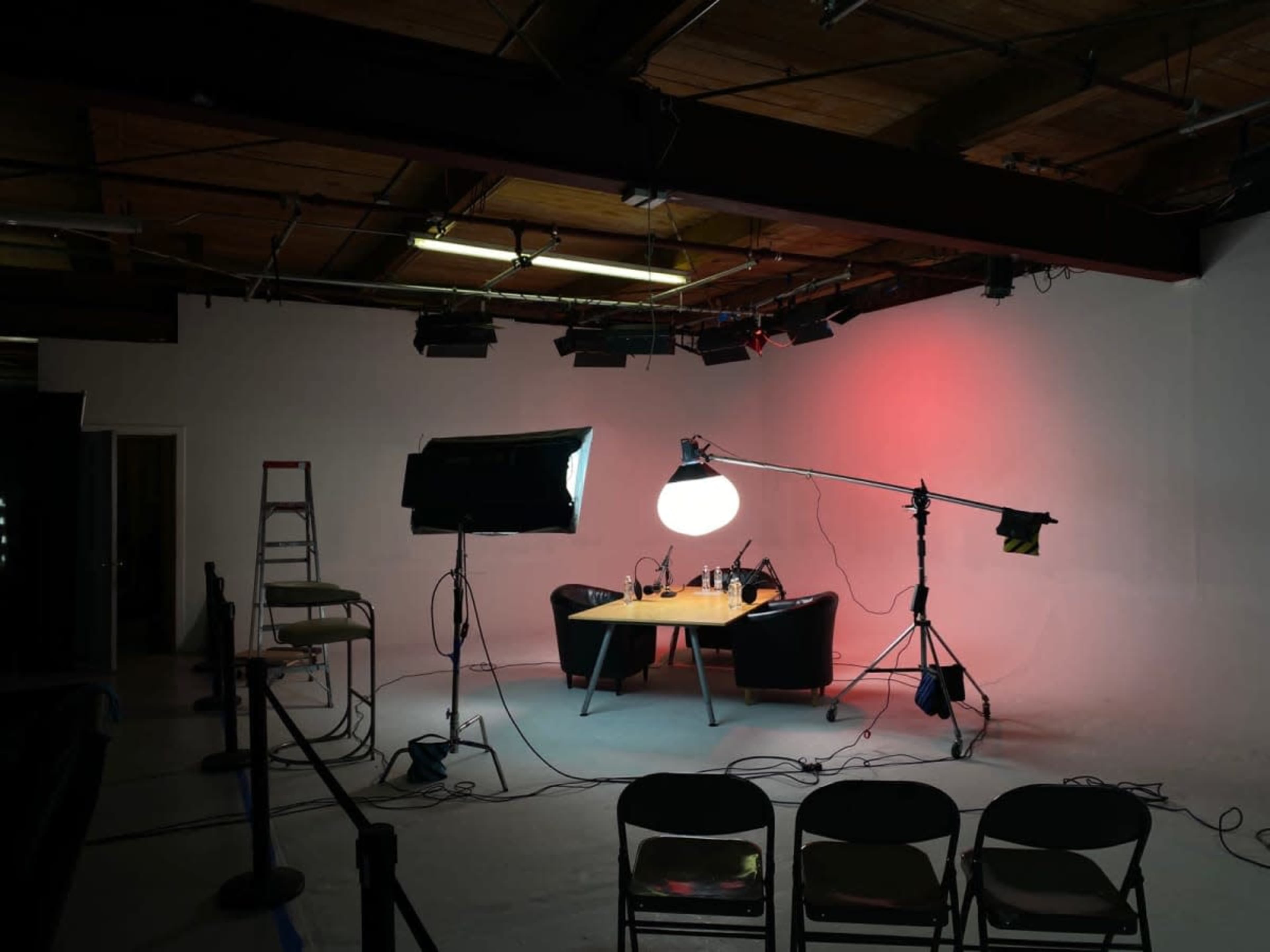 The image shows a minimalist studio setup with two black chairs facing a wooden table, illuminated by overhead lights against a plain white backdrop.