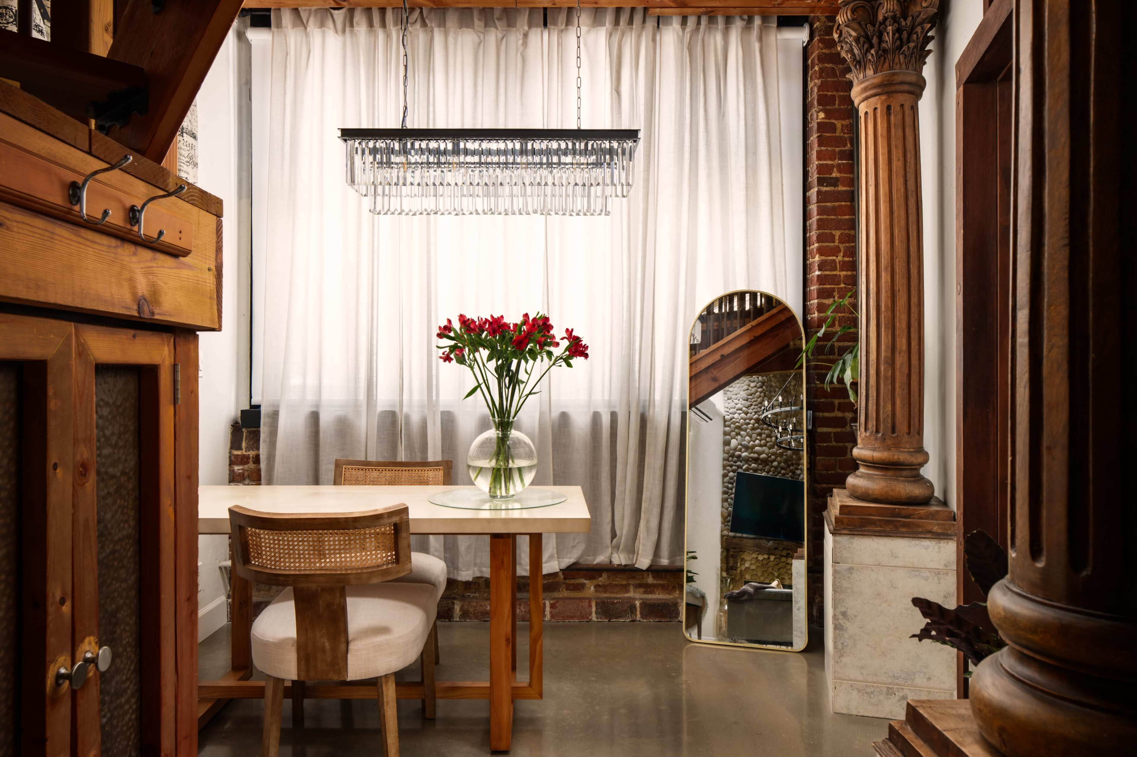 The image depicts a dining area featuring a wooden table with chairs, a large vase of red flowers, a framed mirror, and a chandelier hung above, all set against a backdrop of brick walls and sheer curtains.