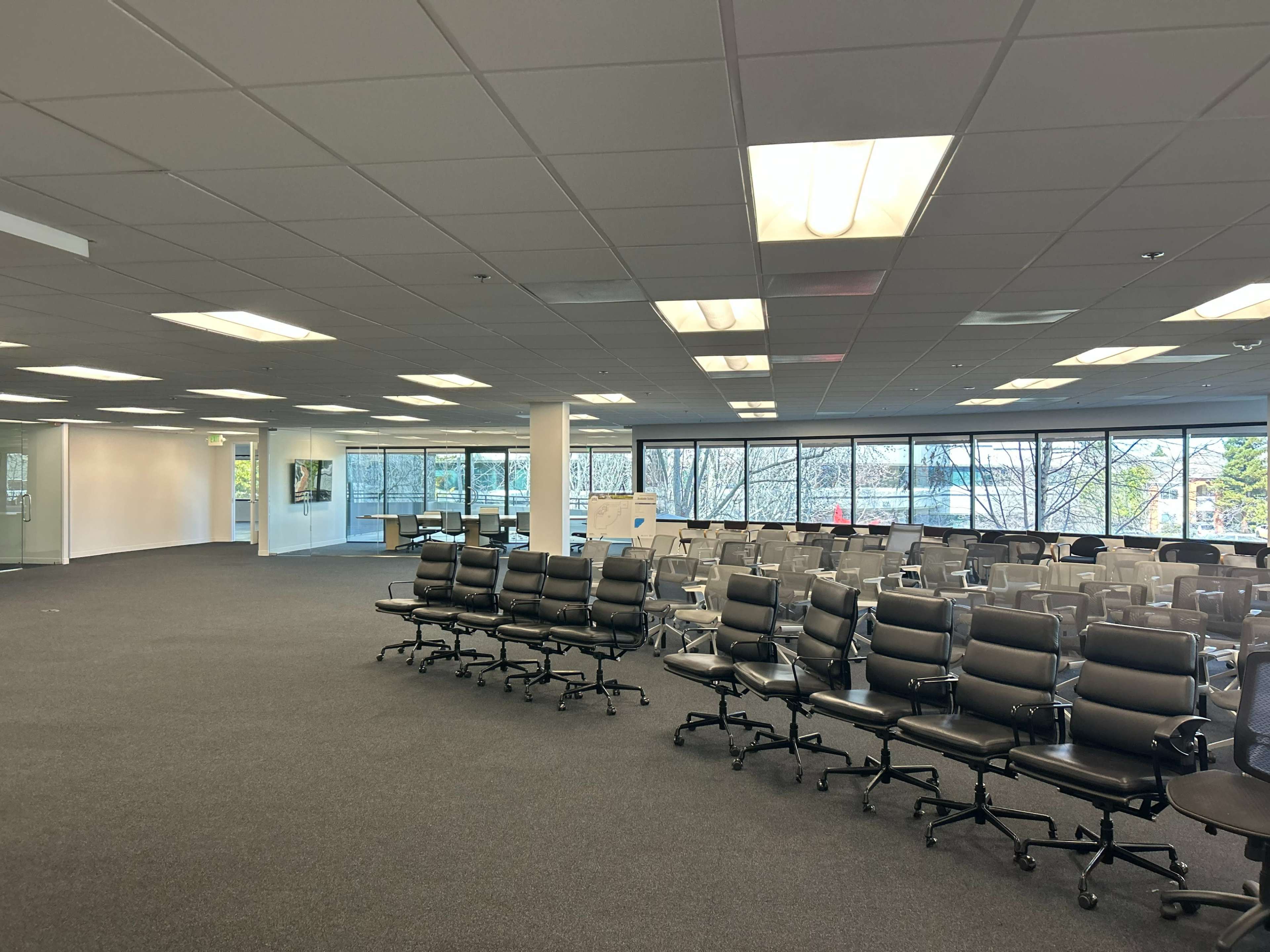 The image shows a large, empty office space with rows of black office chairs set up in front of a wall of windows.