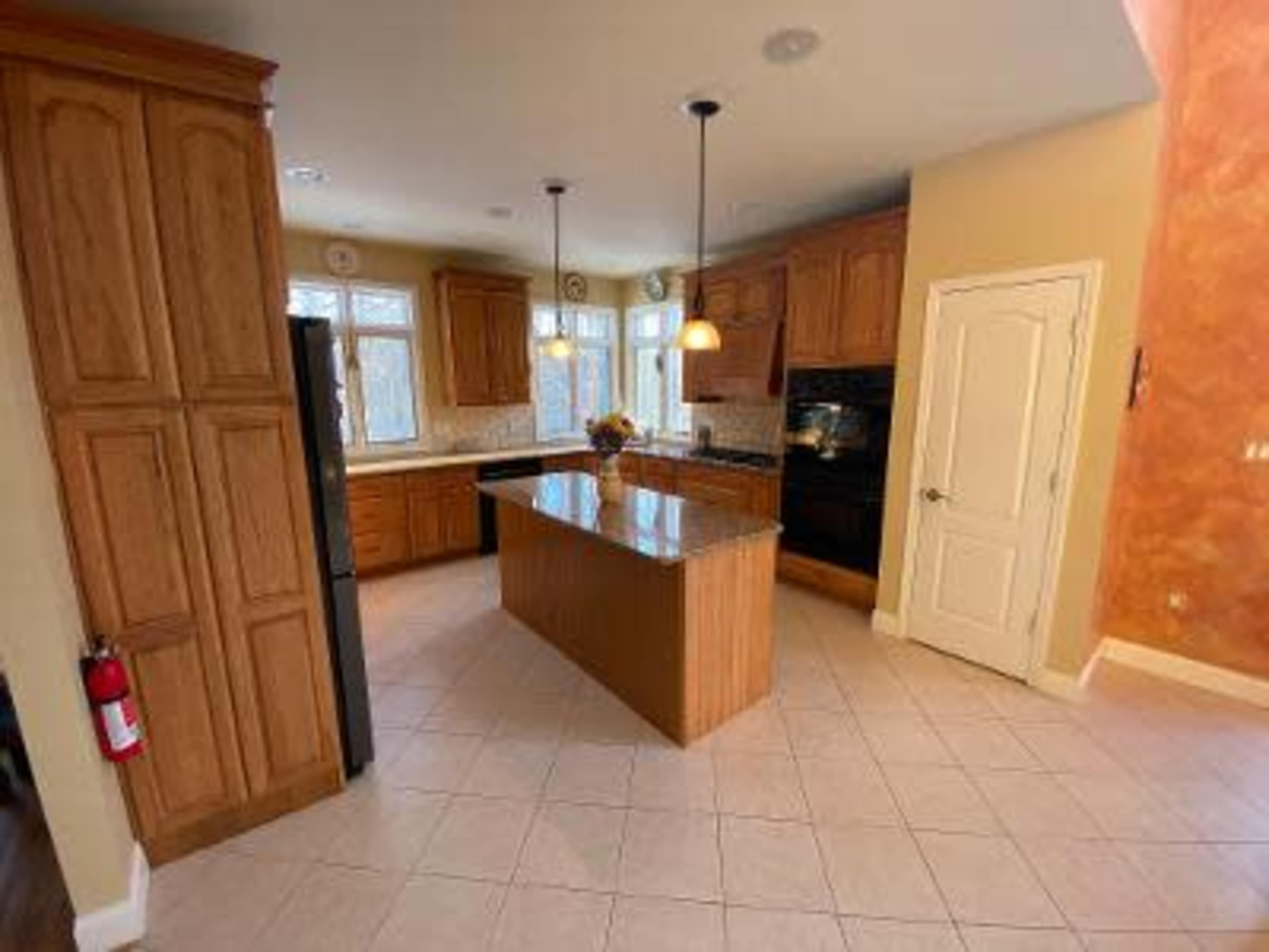 The image shows a spacious kitchen with wooden cabinets, a central island with a granite countertop, and tiled flooring.