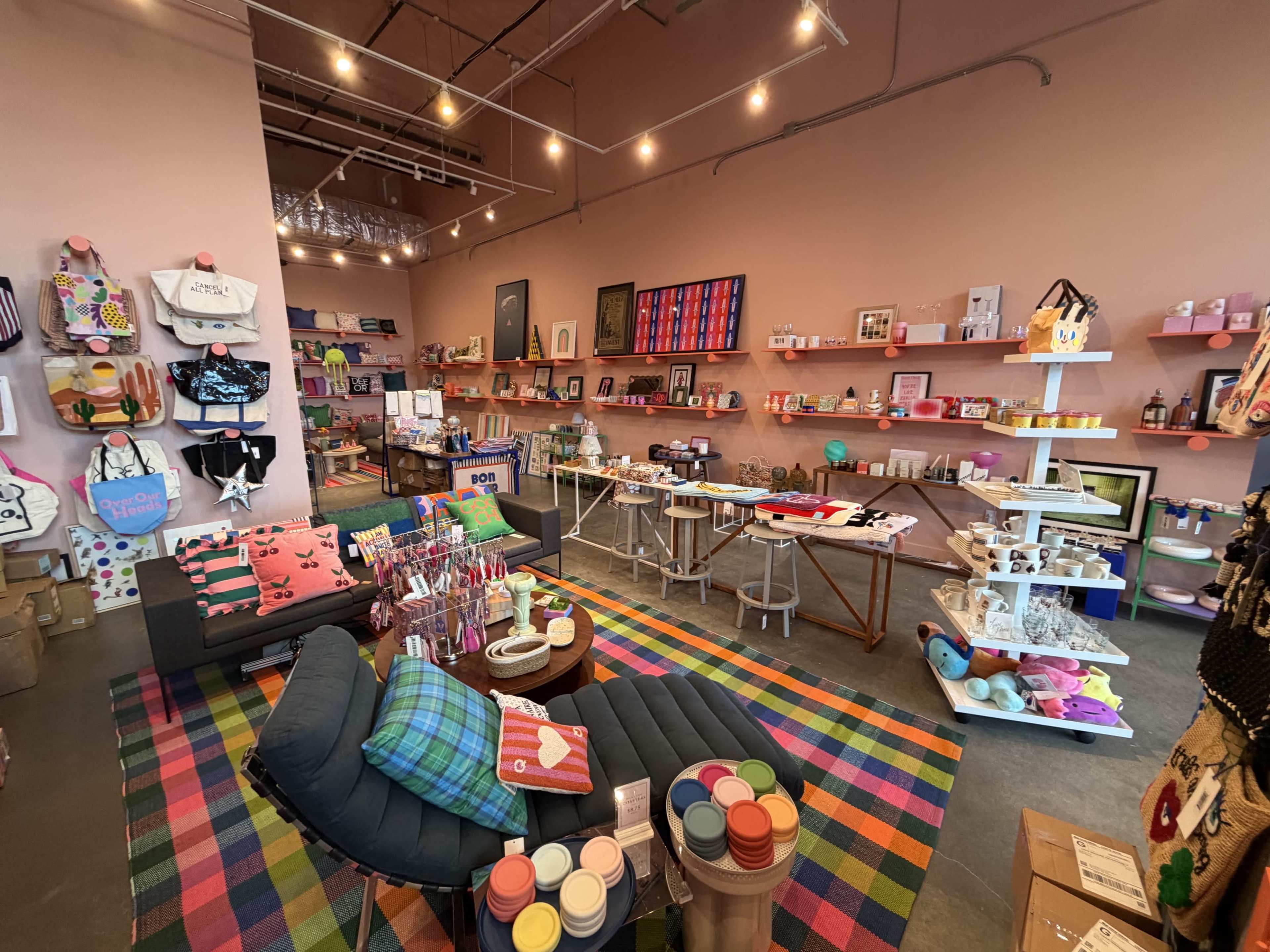 The image shows a colorful retail space filled with various merchandise, including bags, home goods, and beauty products, displayed on shelves against a pink wall.