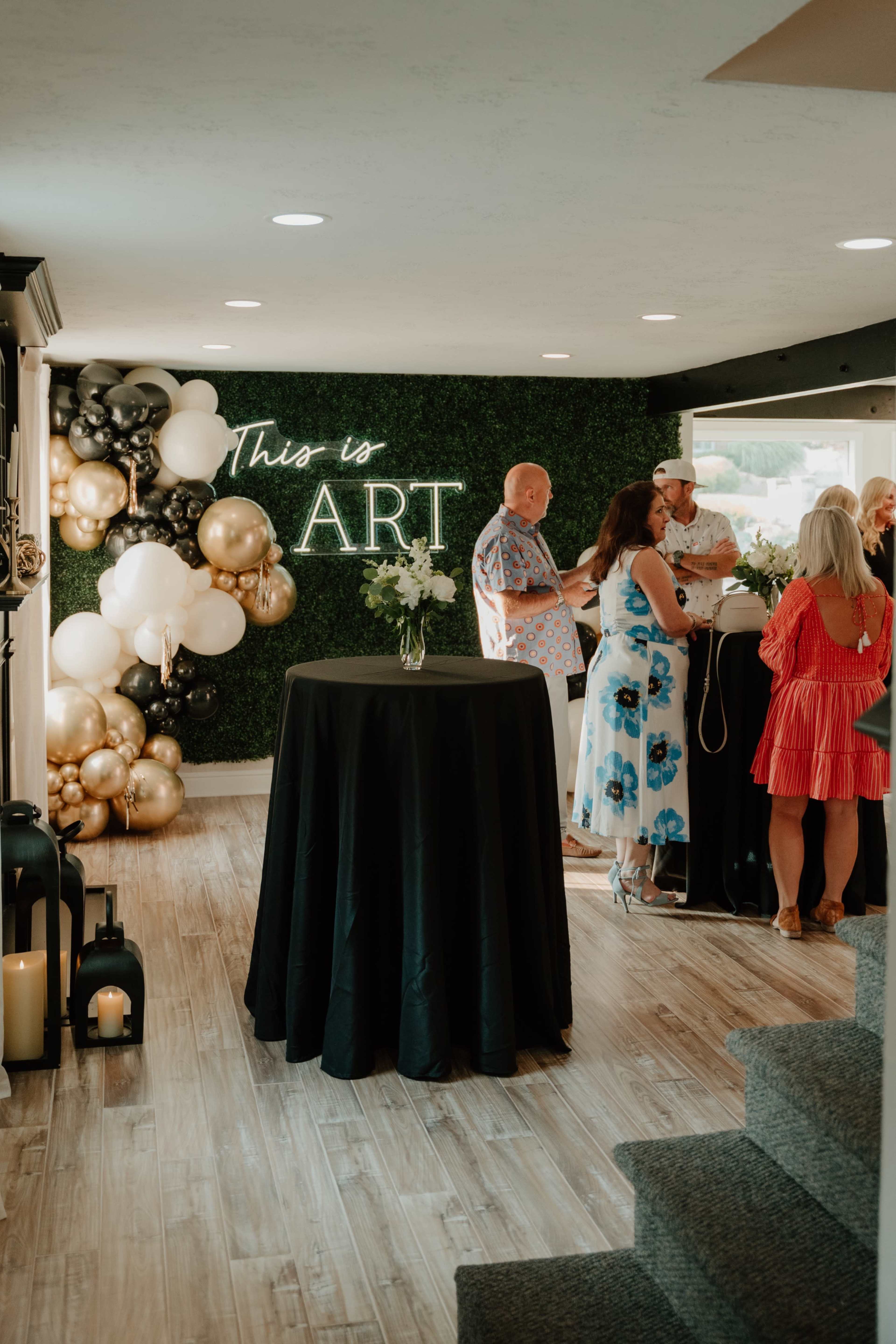 A well-decorated indoor space features a "This is ART" neon sign, a balloon arrangement, and multiple guests mingling around a black table.