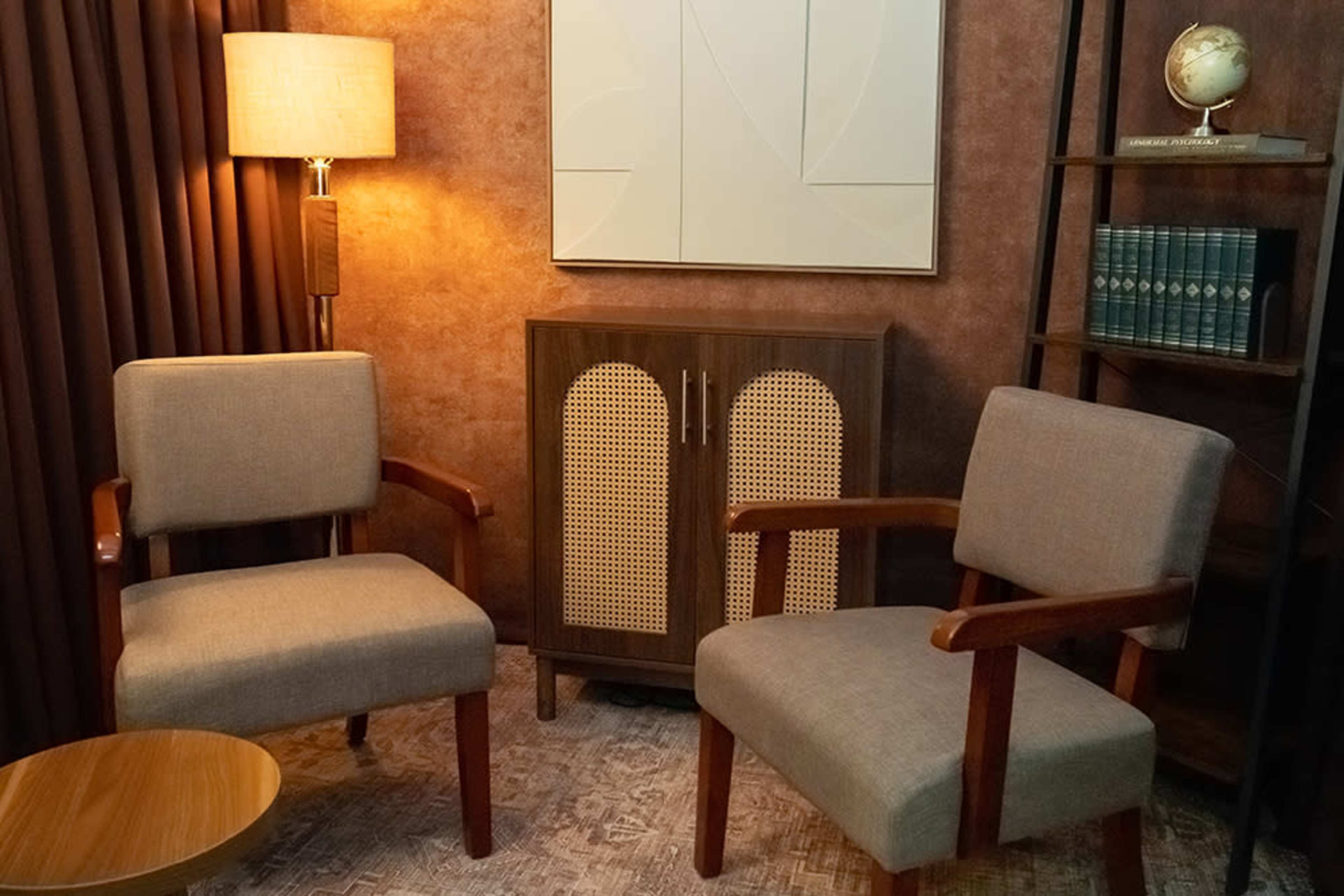 The image shows a cozy room setup featuring two fabric-upholstered chairs, a small round table, a wooden cabinet with cane detail, a bookshelf with books, and warm lighting from a floor lamp.