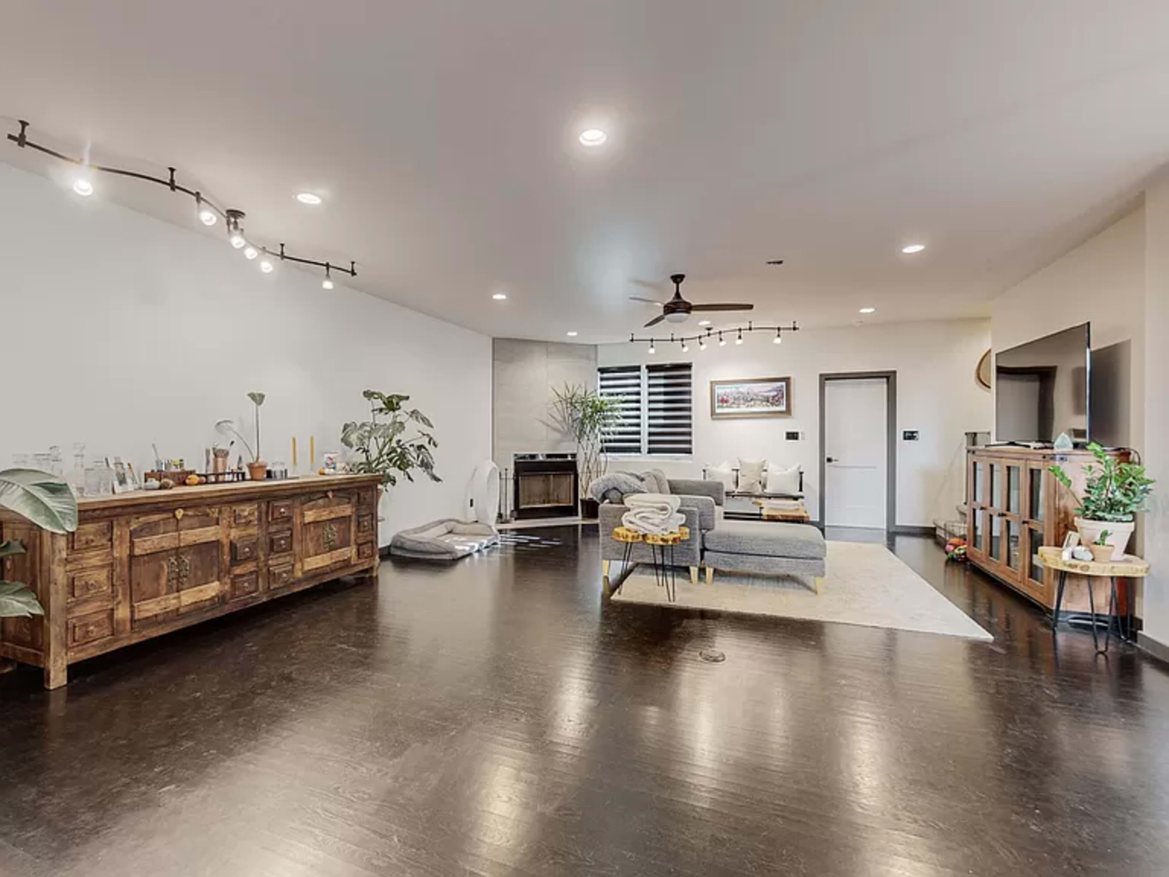 A modern living room featuring a sectional sofa, wooden sideboard, and large flat-screen TV against a backdrop of hardwood flooring and indoor plants.