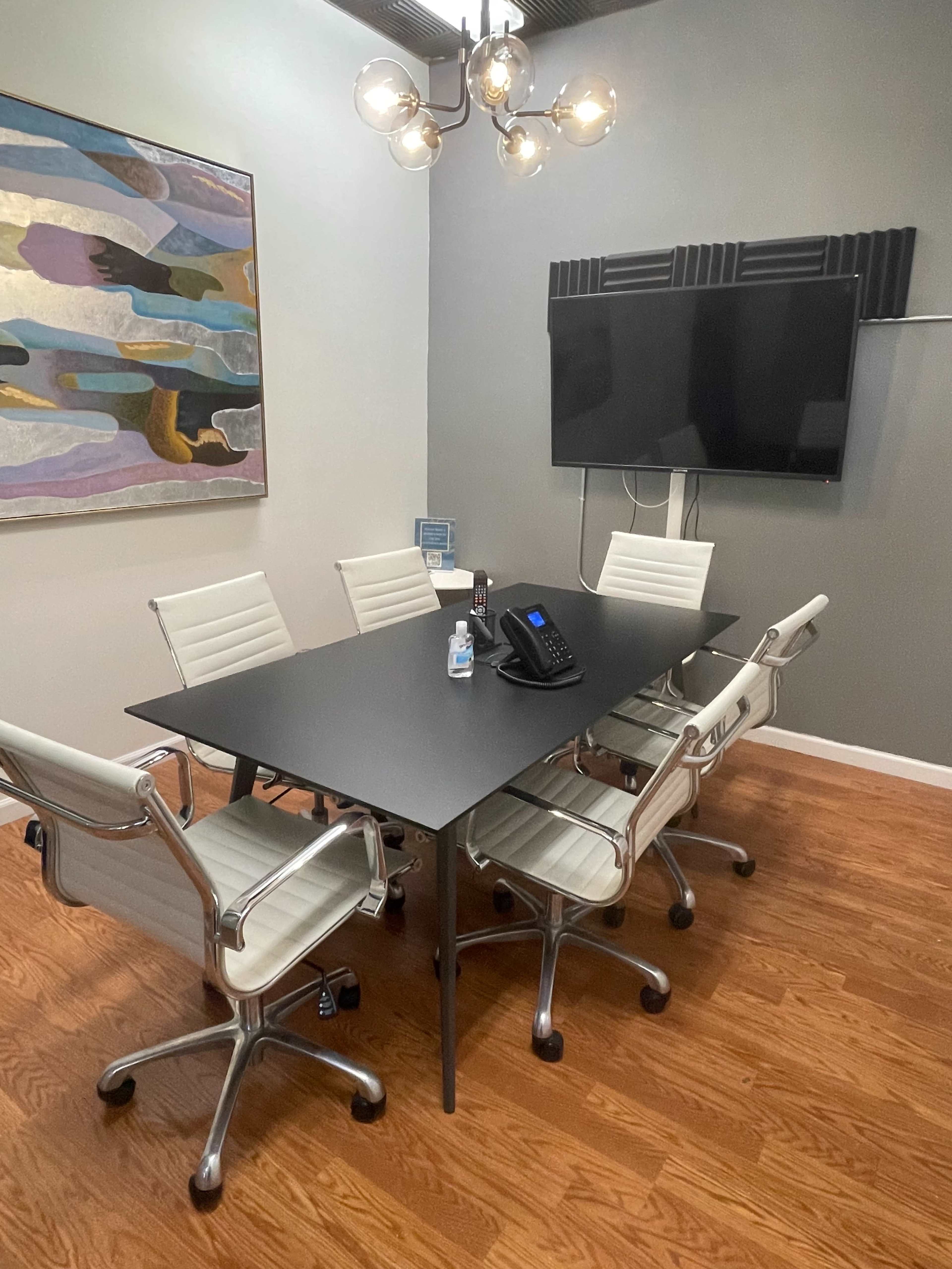 A conference room features a black rectangular table surrounded by five white chairs, a wall-mounted television, and abstract artwork.