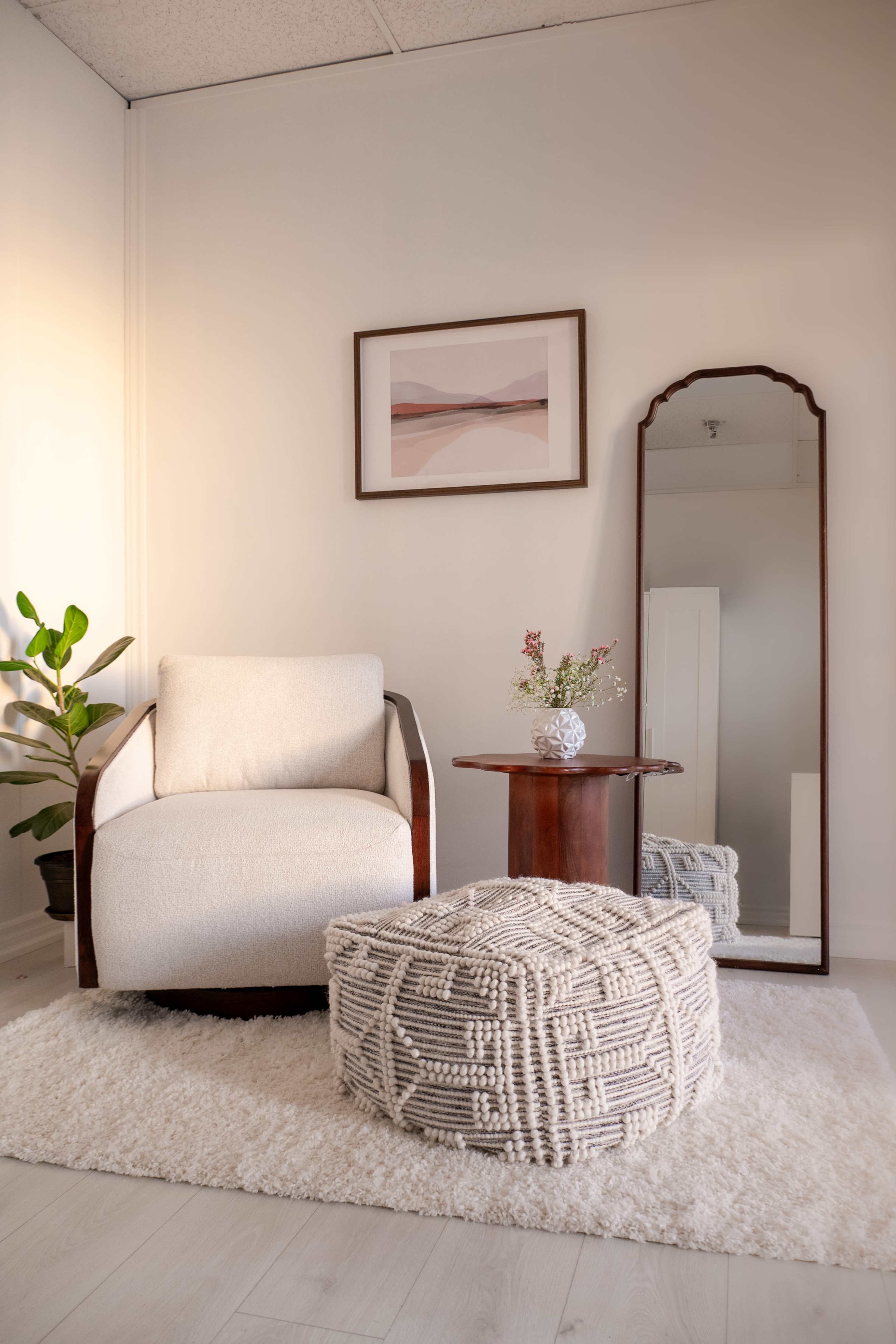 A cozy seating area features a white armchair, a round wooden side table, a patterned pouf, a mirror, and a potted plant on a soft rug.