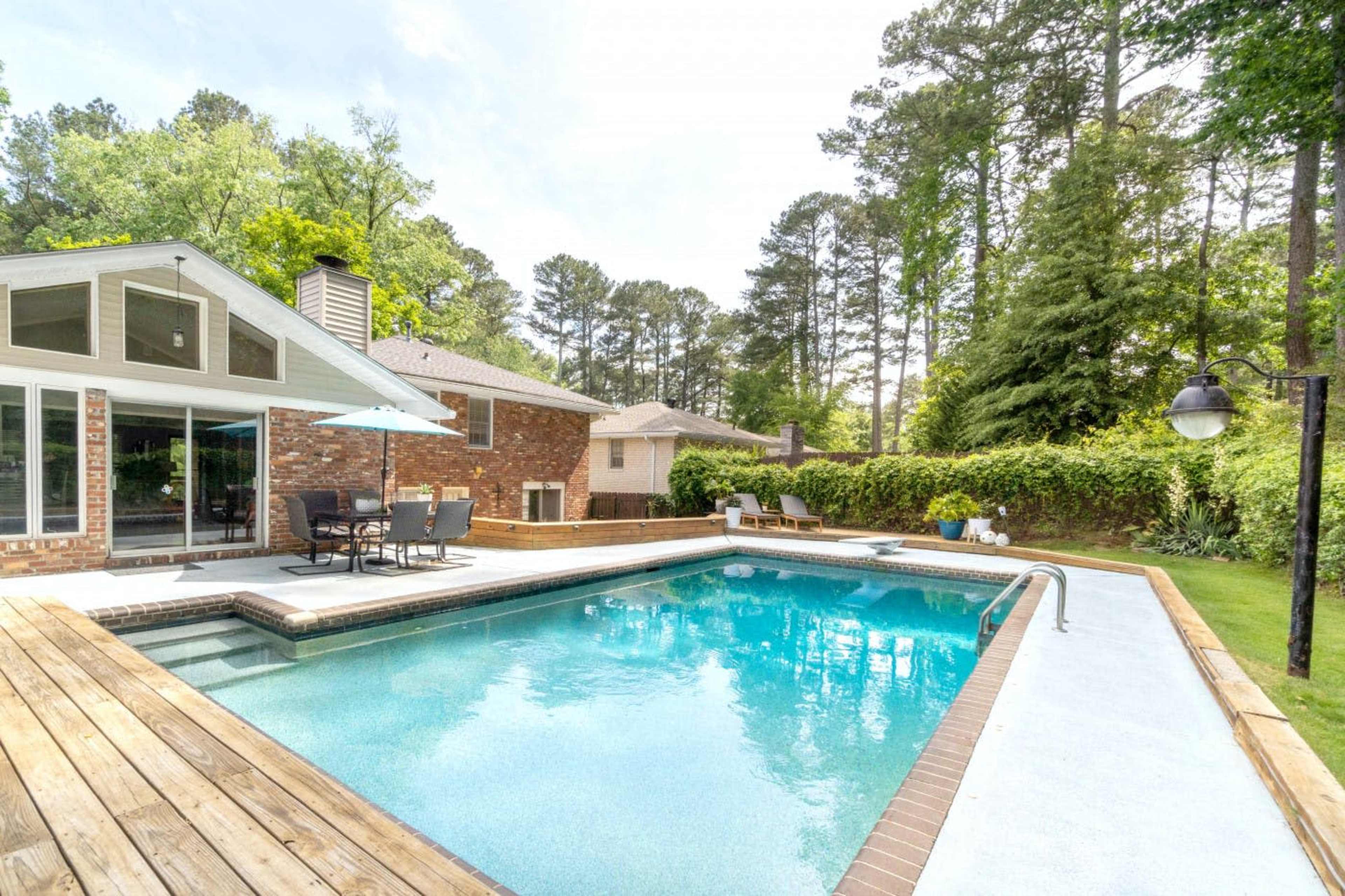 A backyard swimming pool surrounded by wooden decking, with a patio area and tall trees in the background.
