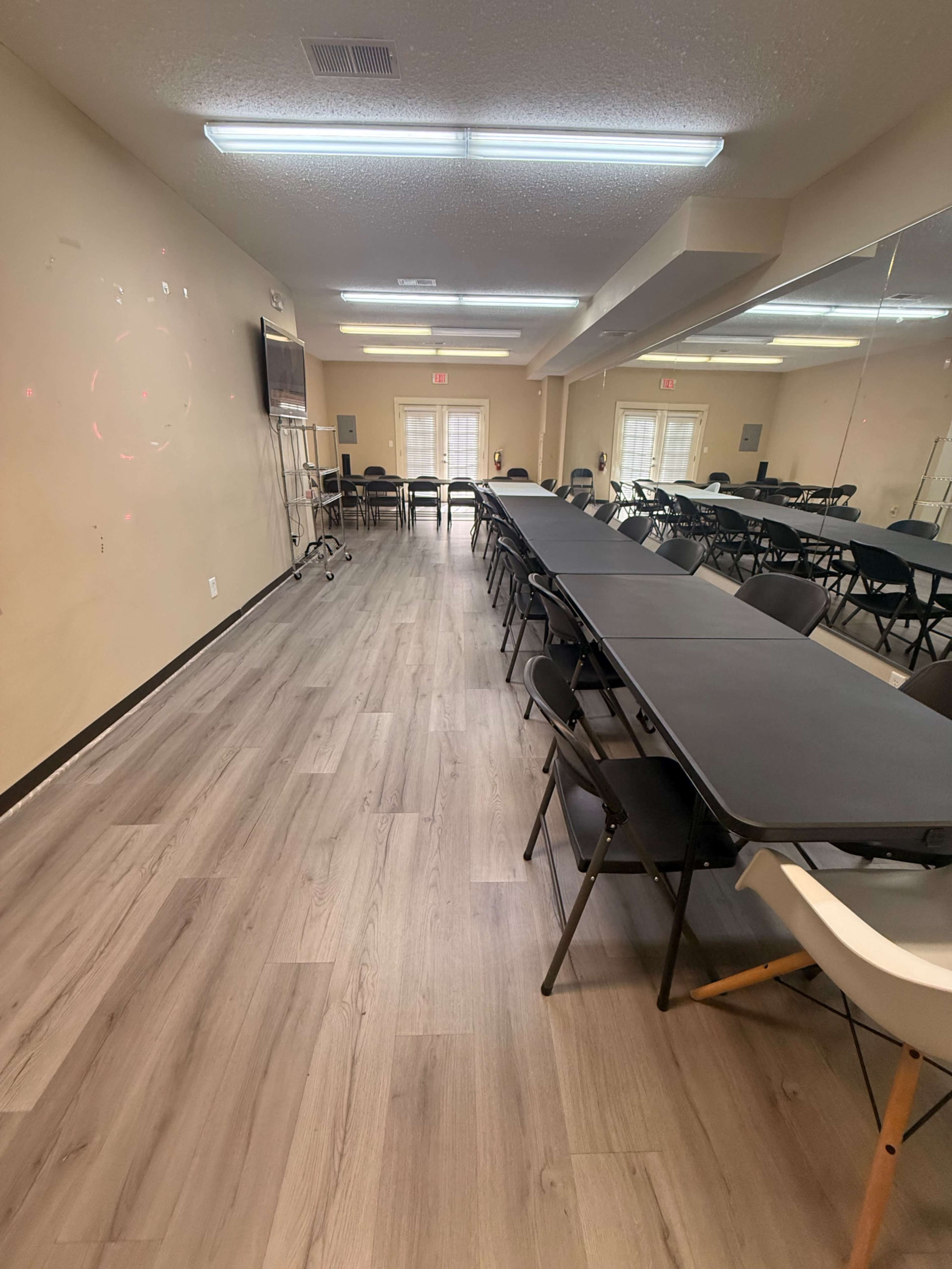 The image shows a spacious room with rows of black folding tables and chairs, featuring a large mirror and a television mounted on the wall.