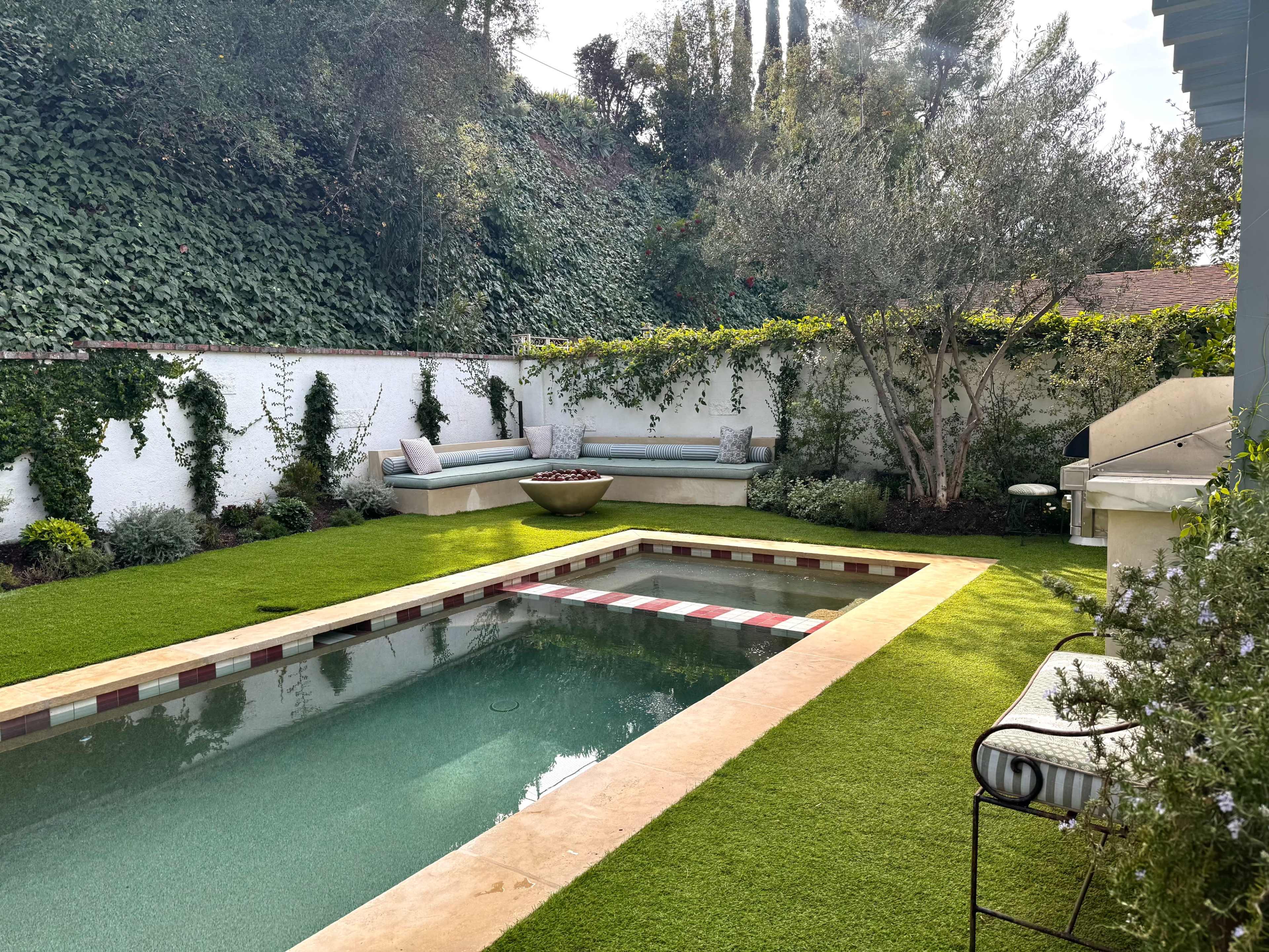 The image shows a backyard with a rectangular swimming pool surrounded by artificial grass, a seating area on one side, and greenery along the walls.