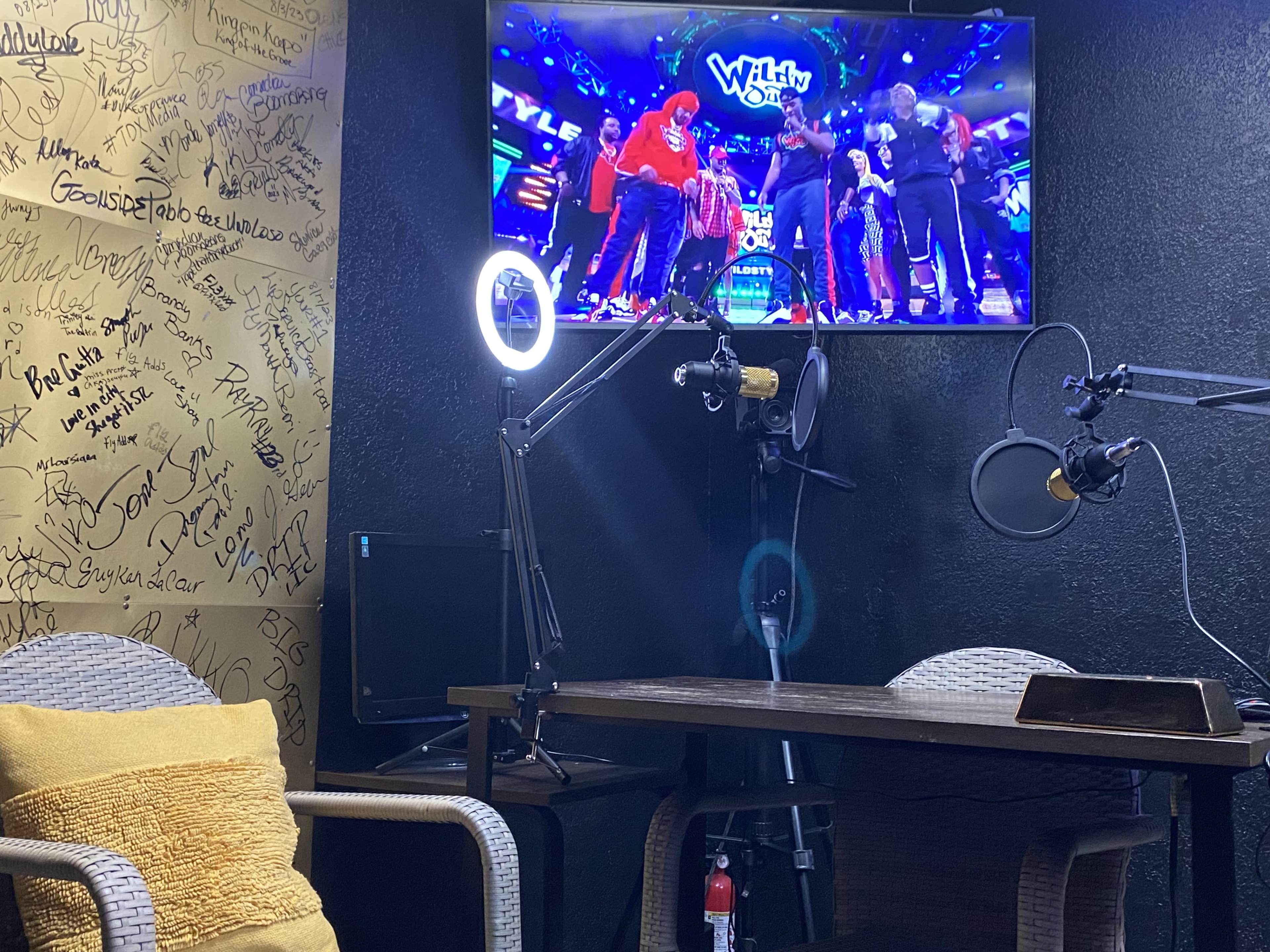 The image shows a well-lit recording studio featuring a table with microphones, a television displaying a performance, and a wall covered in signatures.