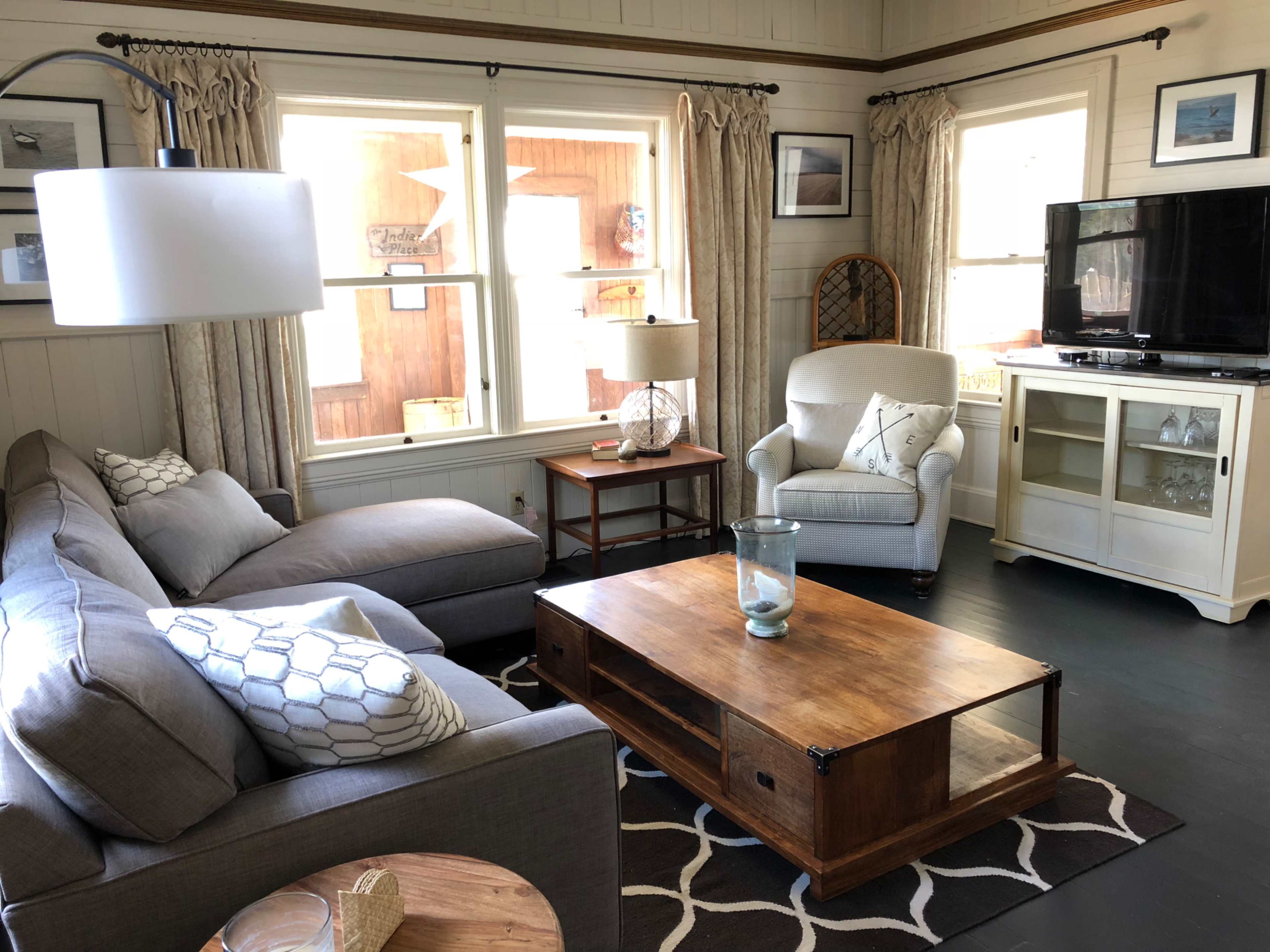 A cozy living room featuring a gray sectional sofa, a light-colored armchair, a wooden coffee table, and large windows.