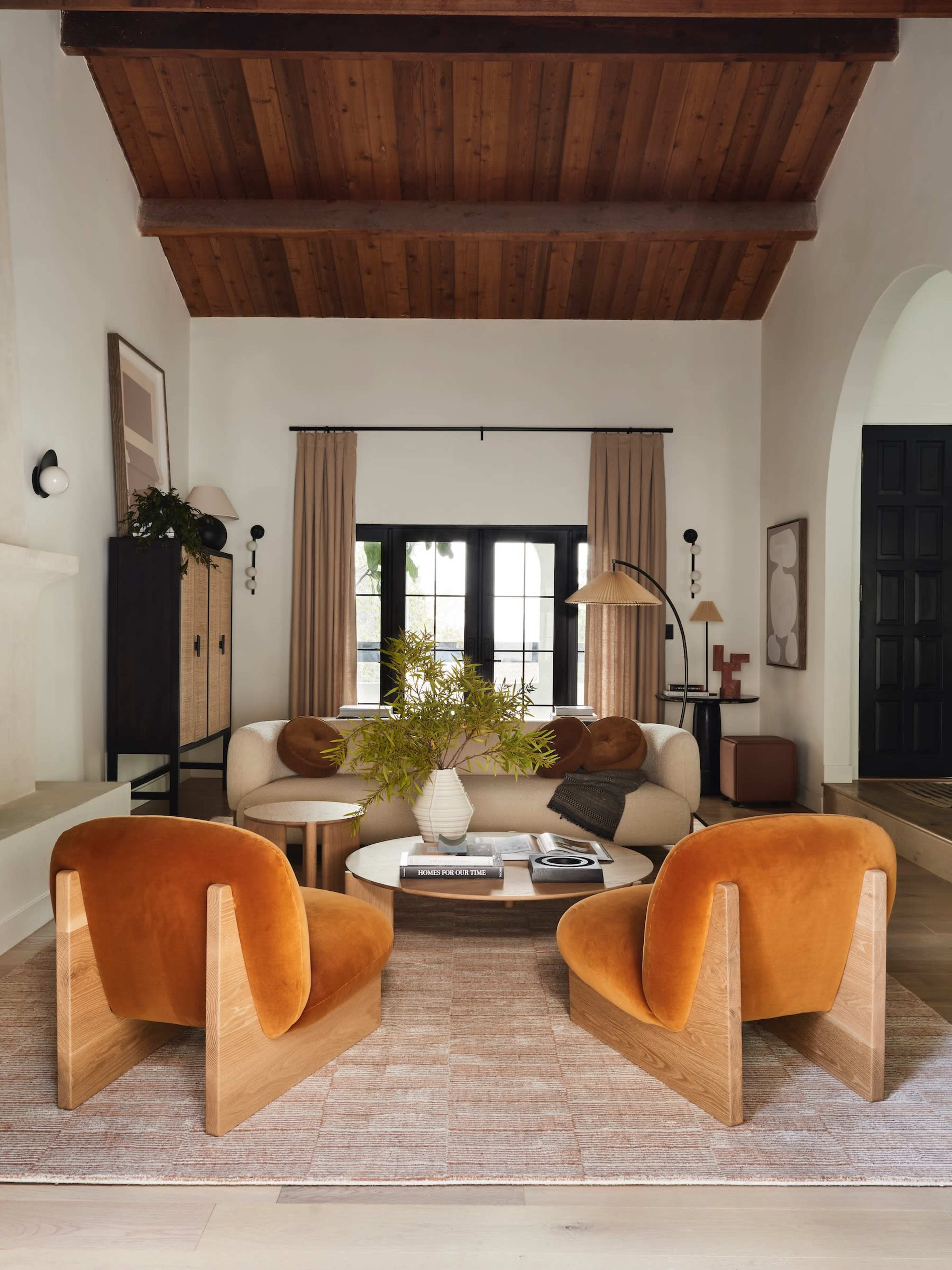 A cozy living room features two orange chairs, a round coffee table, and large windows with light curtains.