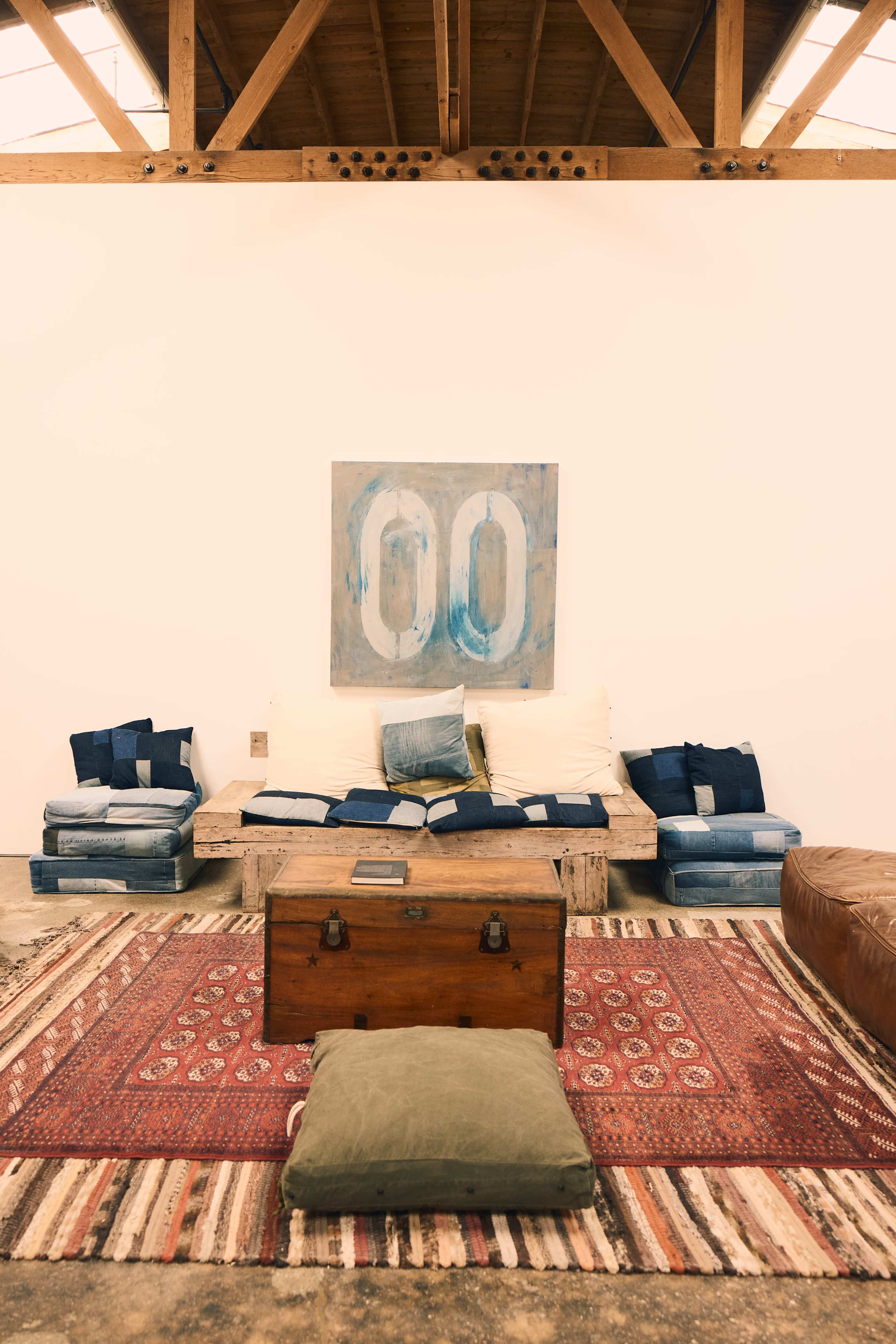 The scene depicts a minimalist living space with a low wooden coffee table, a colorful patterned rug, and a sofa adorned with various blue cushions, all set against a plain white wall with a large abstract painting.
