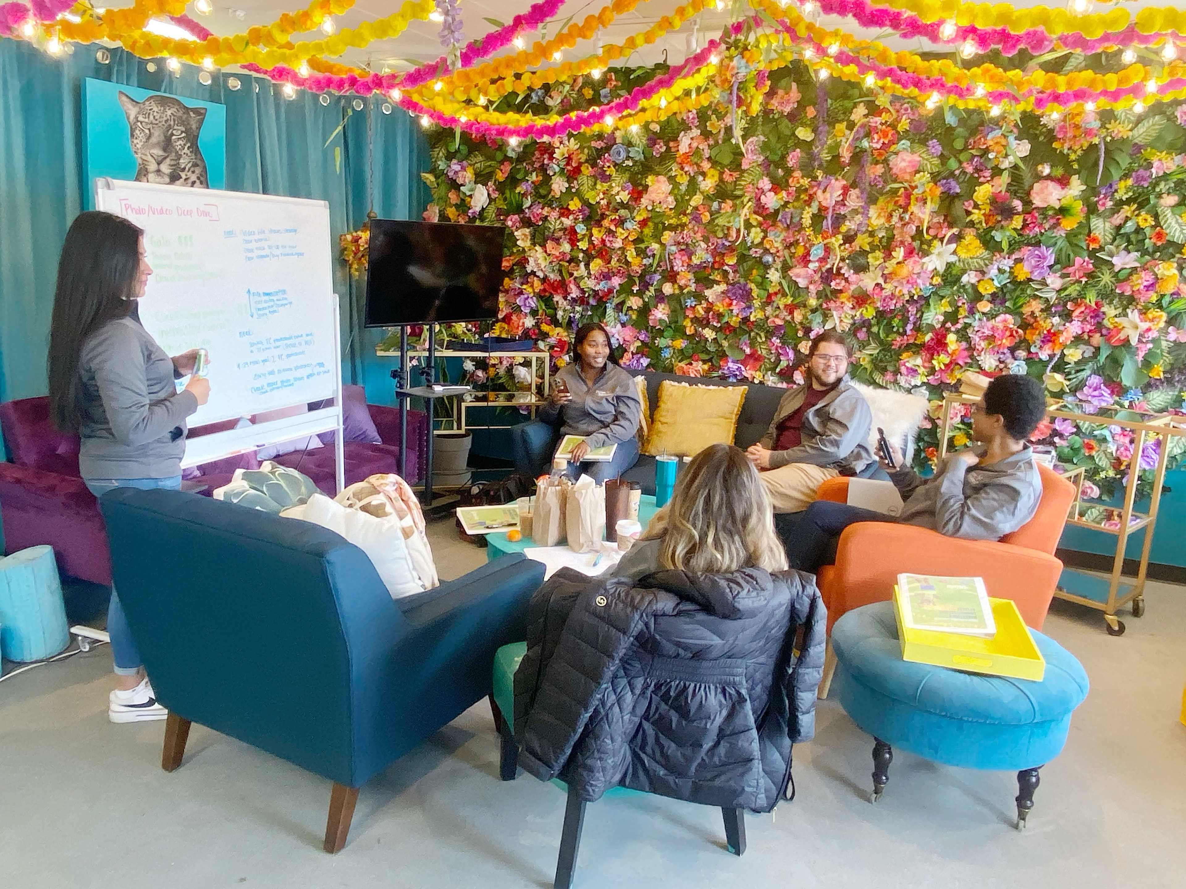 A group of four people is gathered in a colorful, flower-adorned room, engaged in a discussion while seated on various couches and chairs.