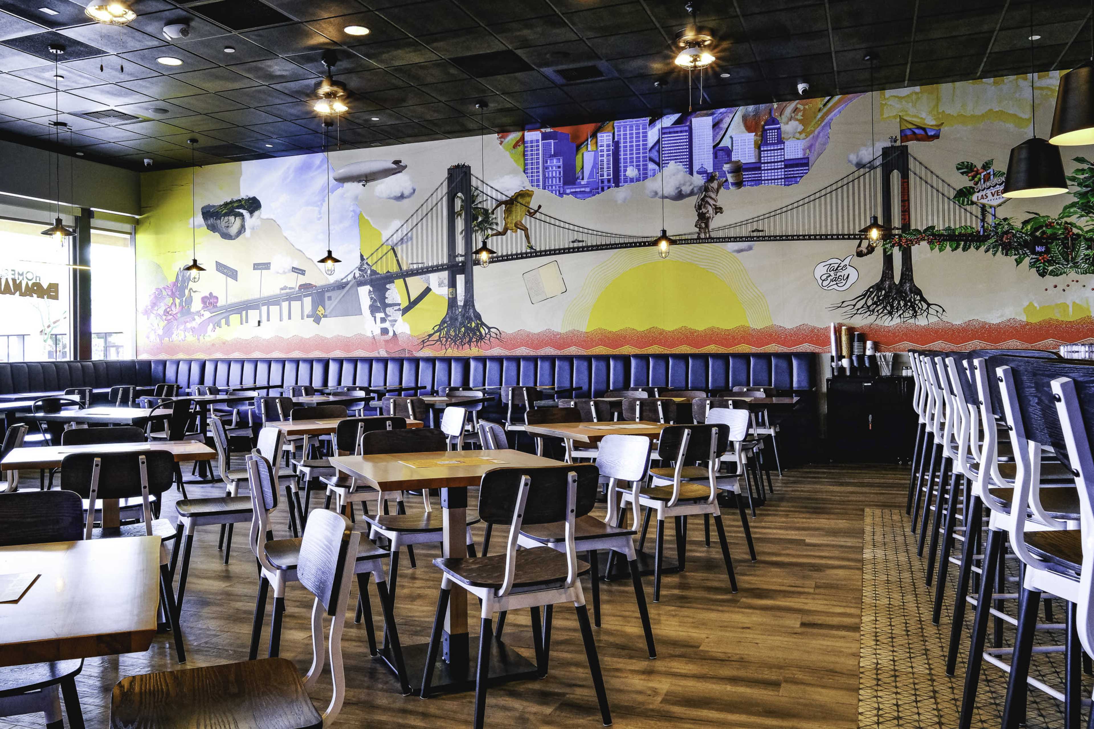 The image shows a modern restaurant interior featuring a mural of a cityscape and bridge on one wall, with rows of empty wooden and metal tables and chairs.
