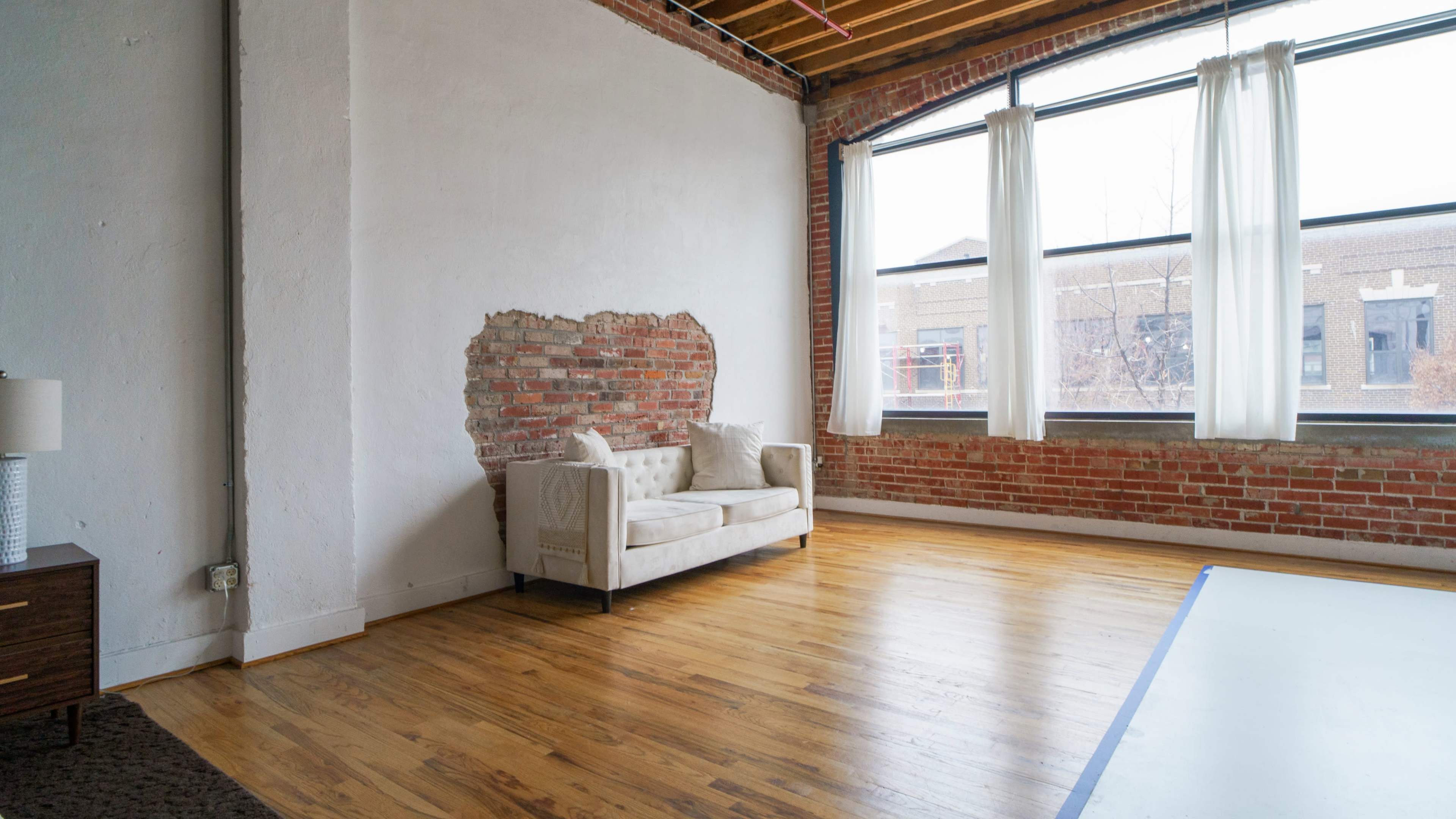 A spacious room with exposed brick walls, a wooden floor, and a light-colored couch near a large window with sheer curtains.
