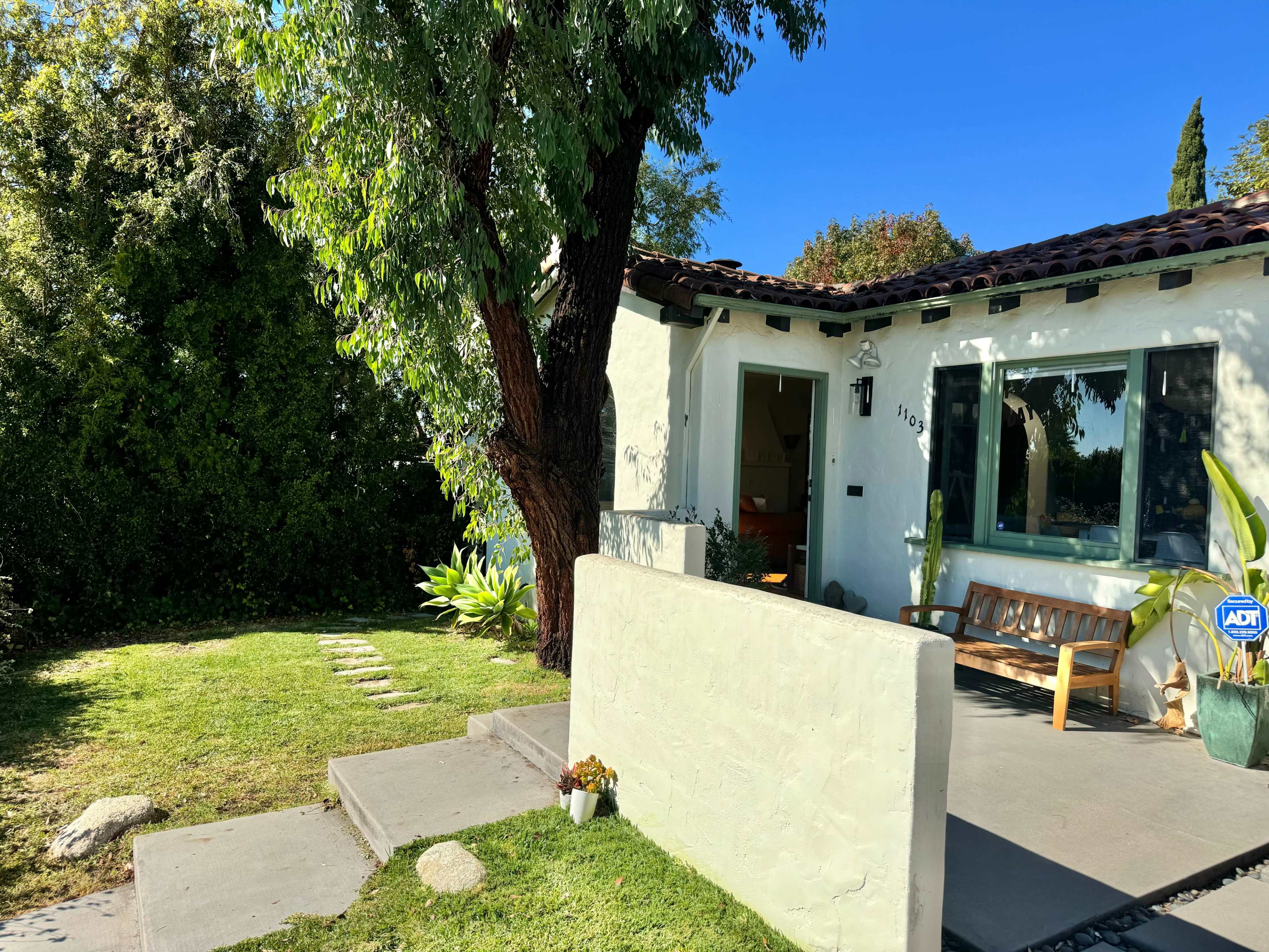 A single-story house with a tiled roof, a front bench, and lush greenery in the yard.