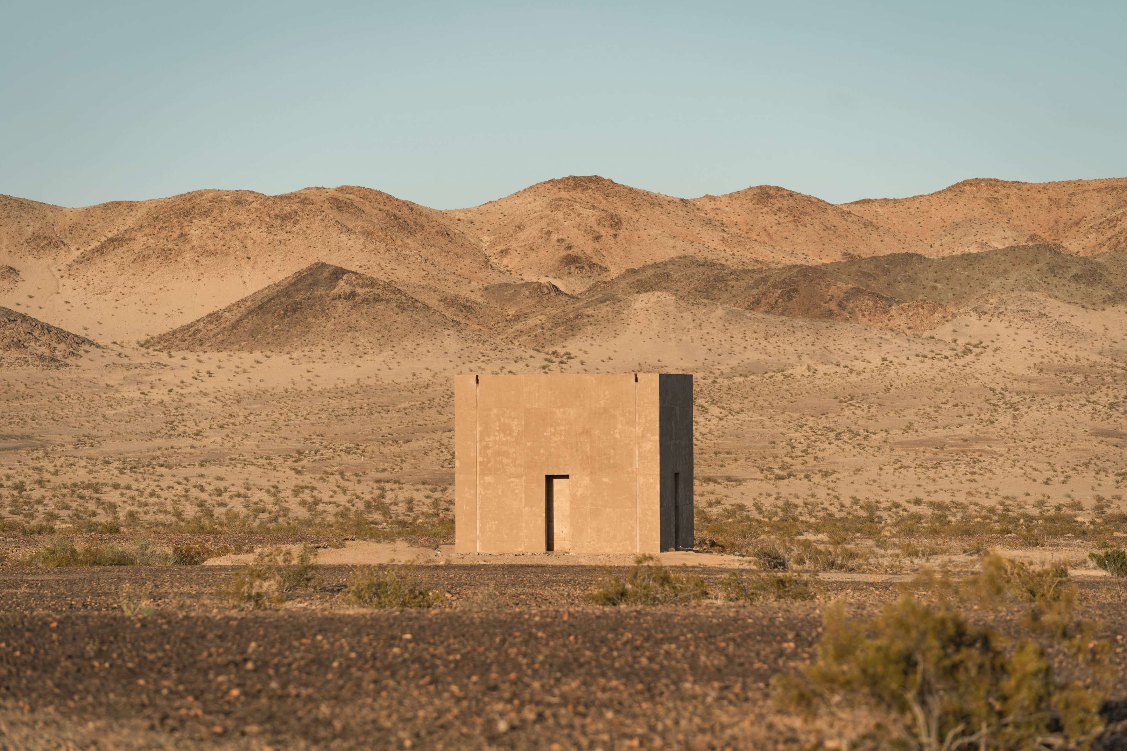A simple, square building stands alone on a barren landscape surrounded by mountains and desert.