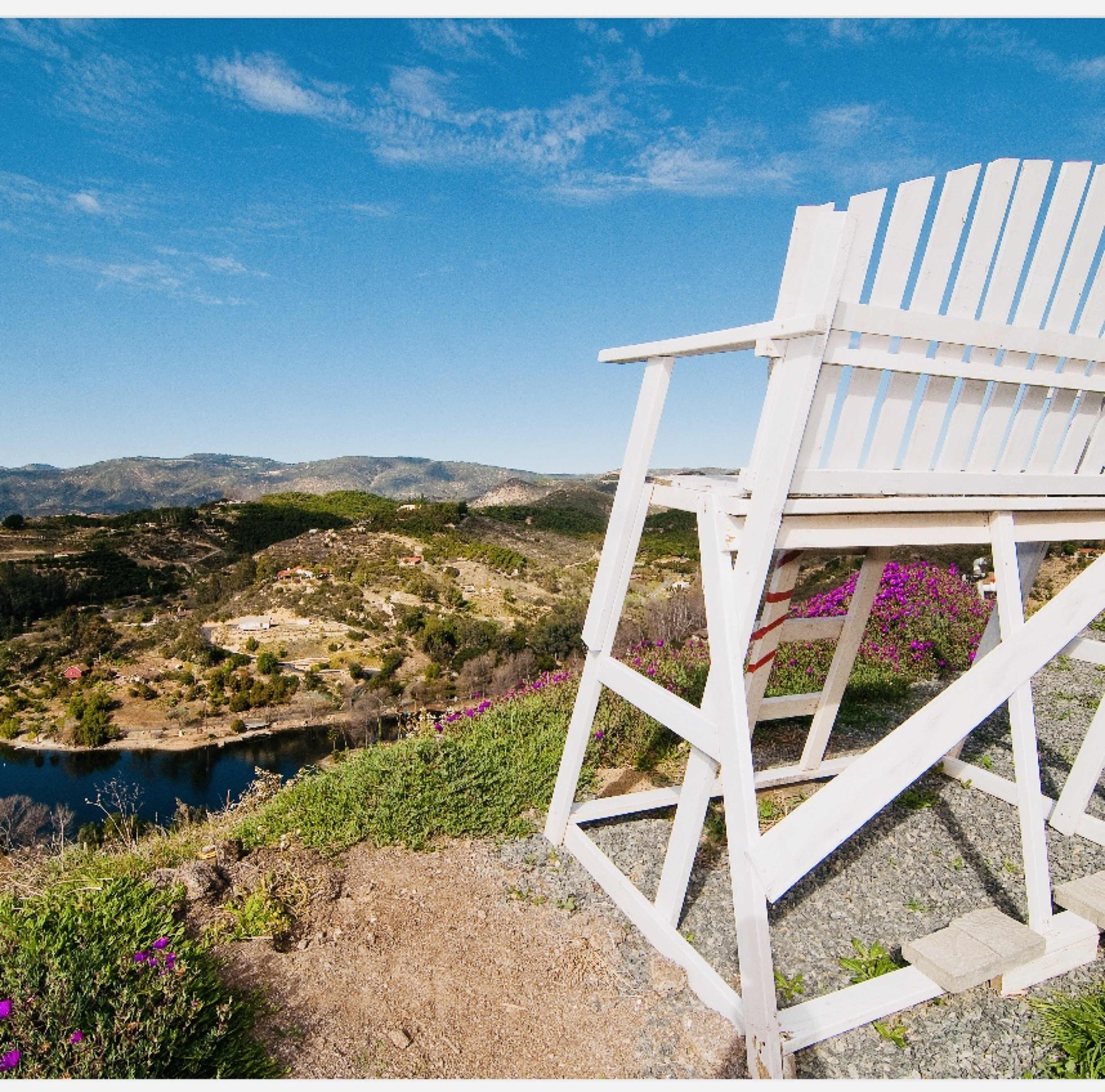A wooden observation chair sits on a hillside overlooking a lush landscape and a tranquil body of water.
