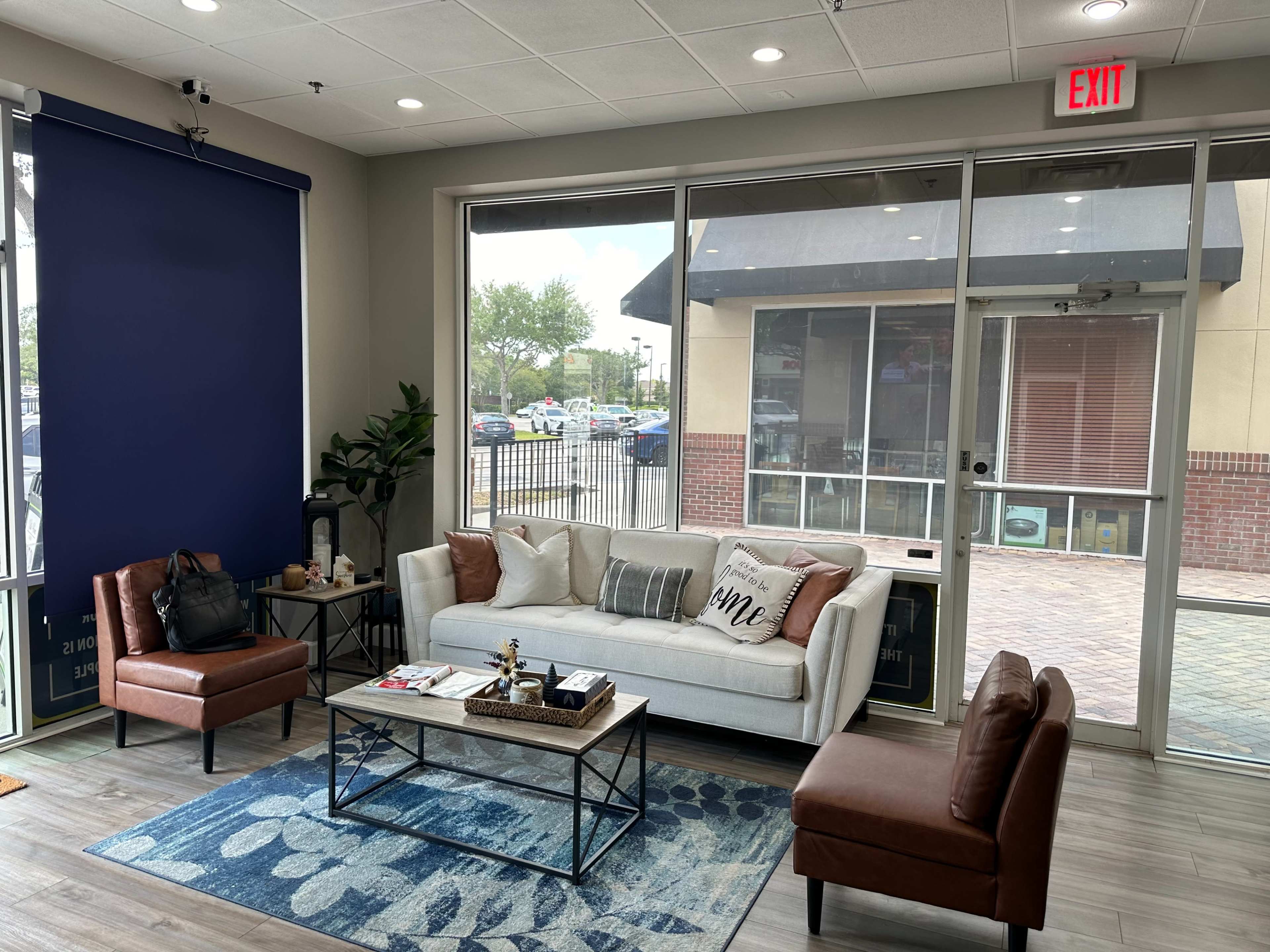 The image shows a modern waiting area with a white couch, two brown leather chairs, a coffee table, and large windows overlooking an outdoor space.