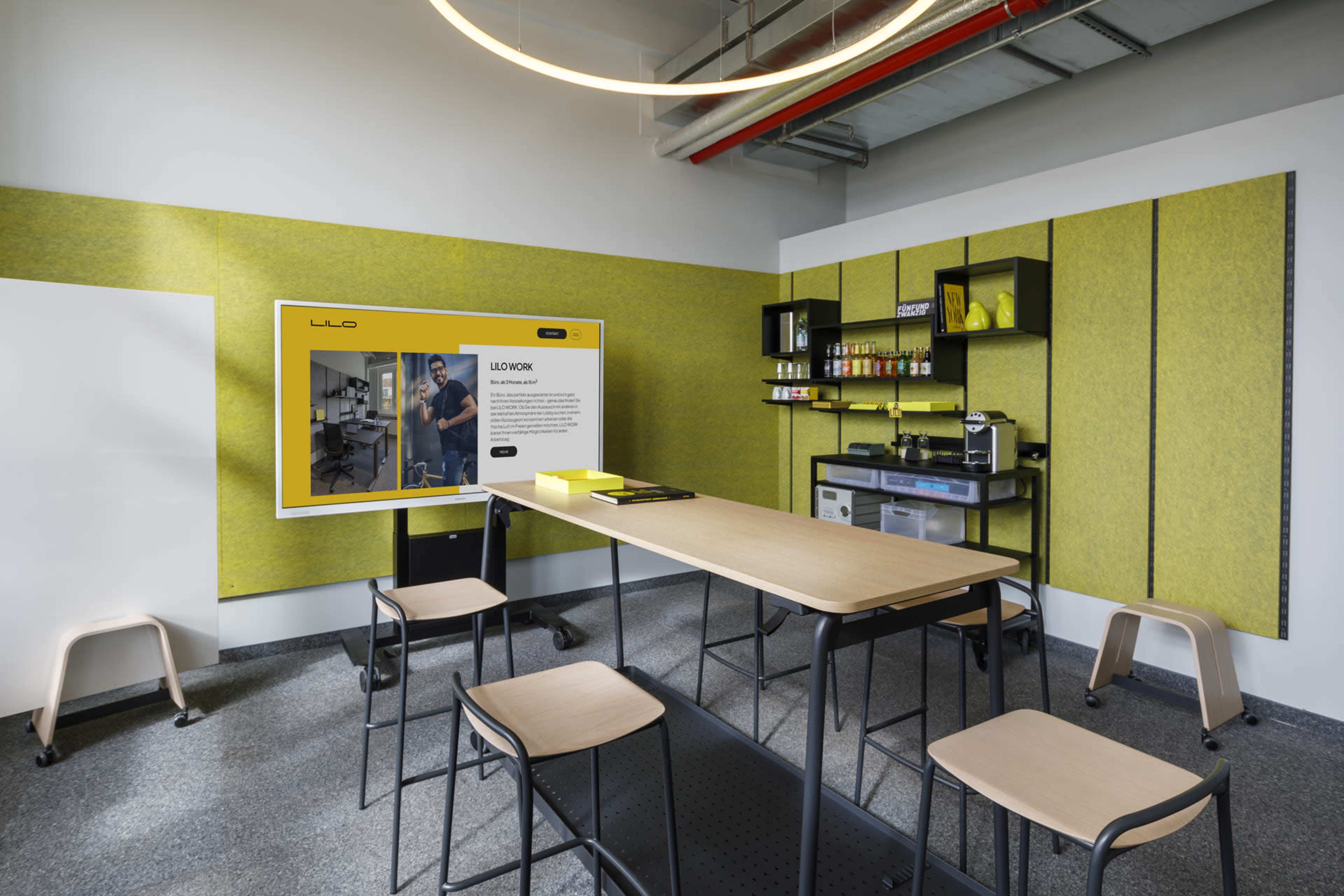 The image shows a modern meeting room with a long table, chairs, a wall-mounted screen displaying information, and a functional kitchenette area.