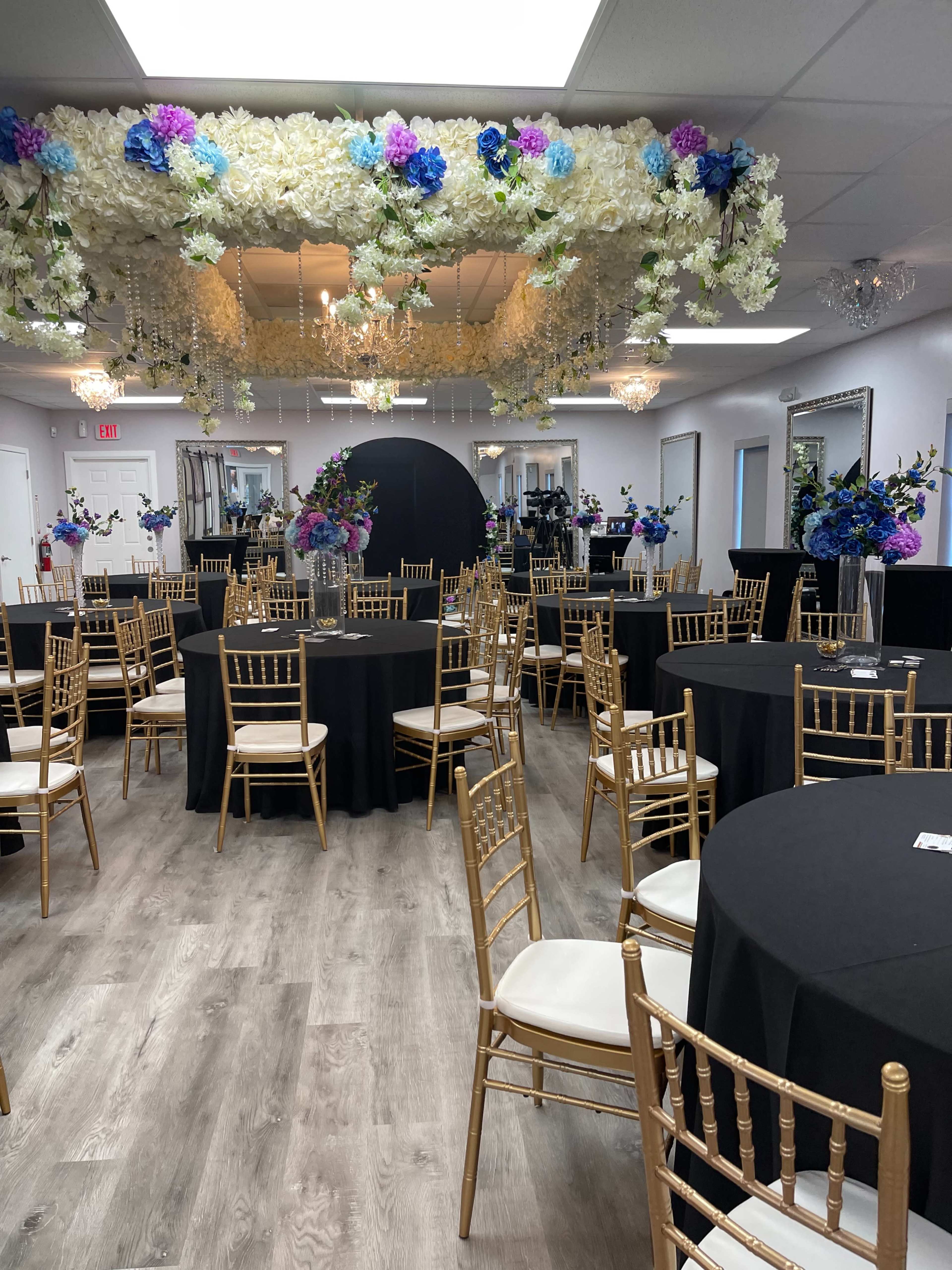 A banquet hall is set up with black tables and gold chairs, adorned with floral arrangements hanging from the ceiling.