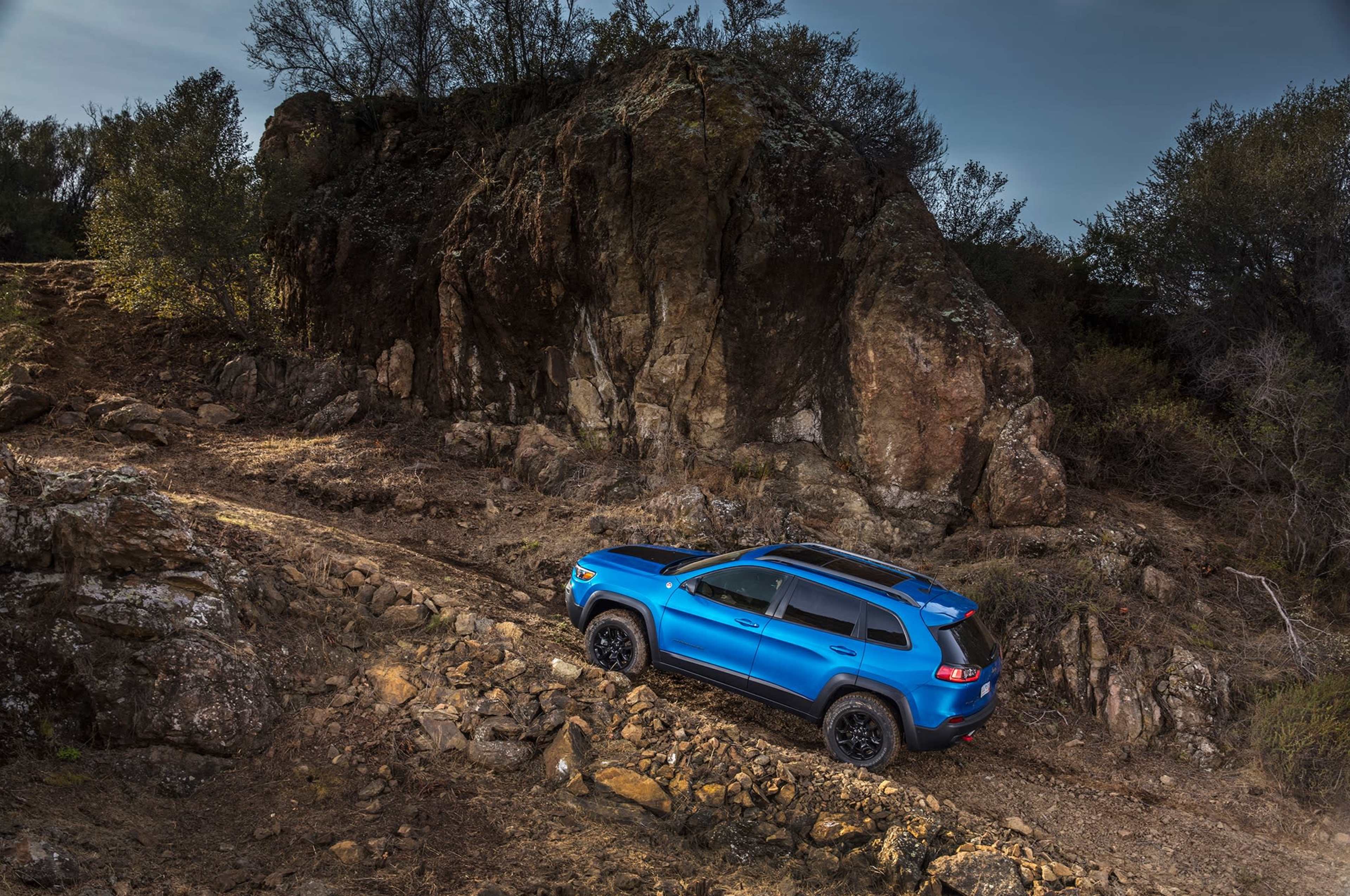A blue SUV ascends a rocky path beside a large boulder in a natural landscape.