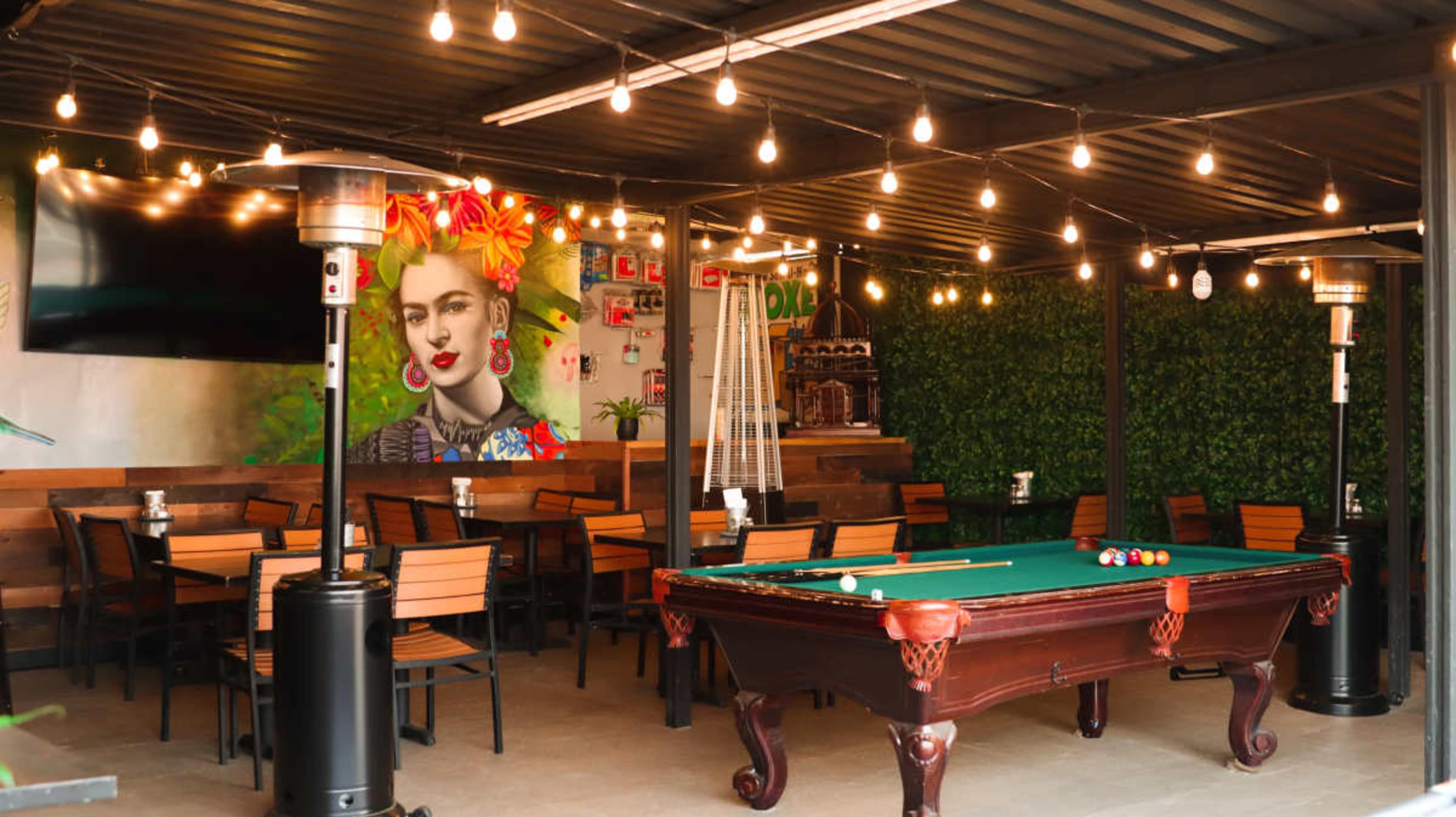 The scene features a vibrant outdoor dining area with a pool table, string lights, and a mural of a woman against a green backdrop.