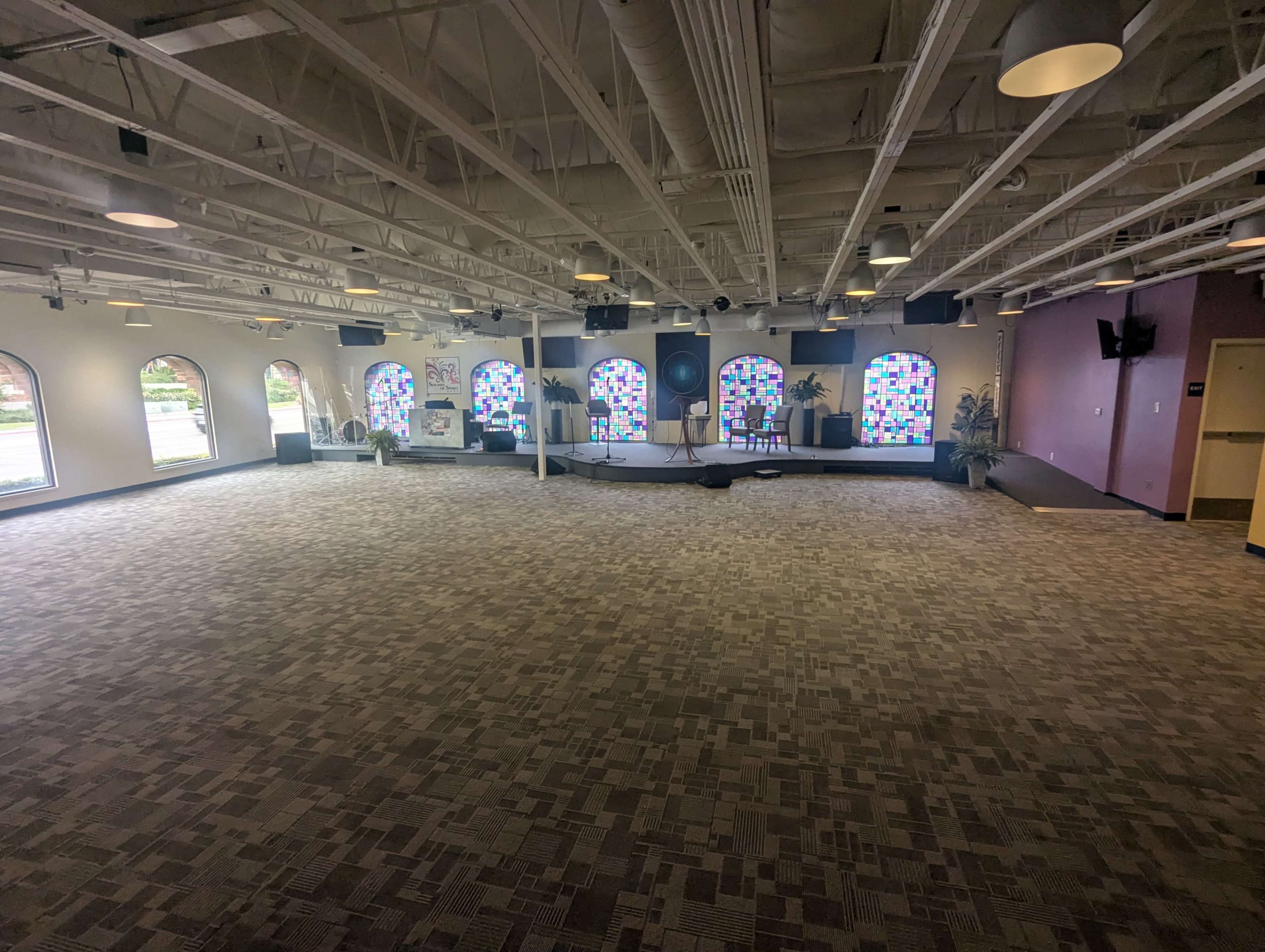 The image shows a spacious, empty room with patterned carpeting, large windows featuring colorful panes, and a stage area at the far end.