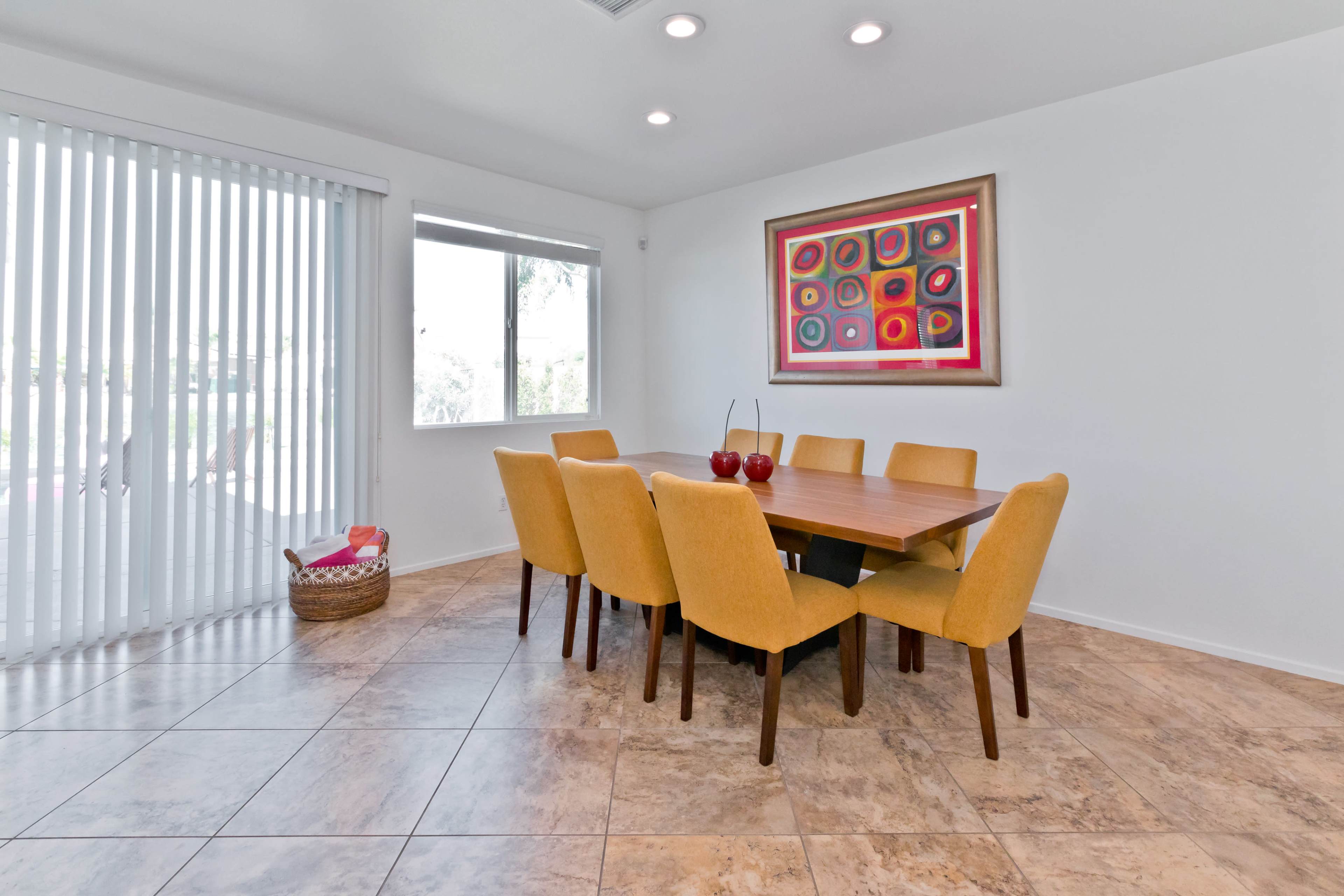 A dining area features a wooden table surrounded by eight yellow chairs, with a large abstract art piece on the wall and a basket in the corner.