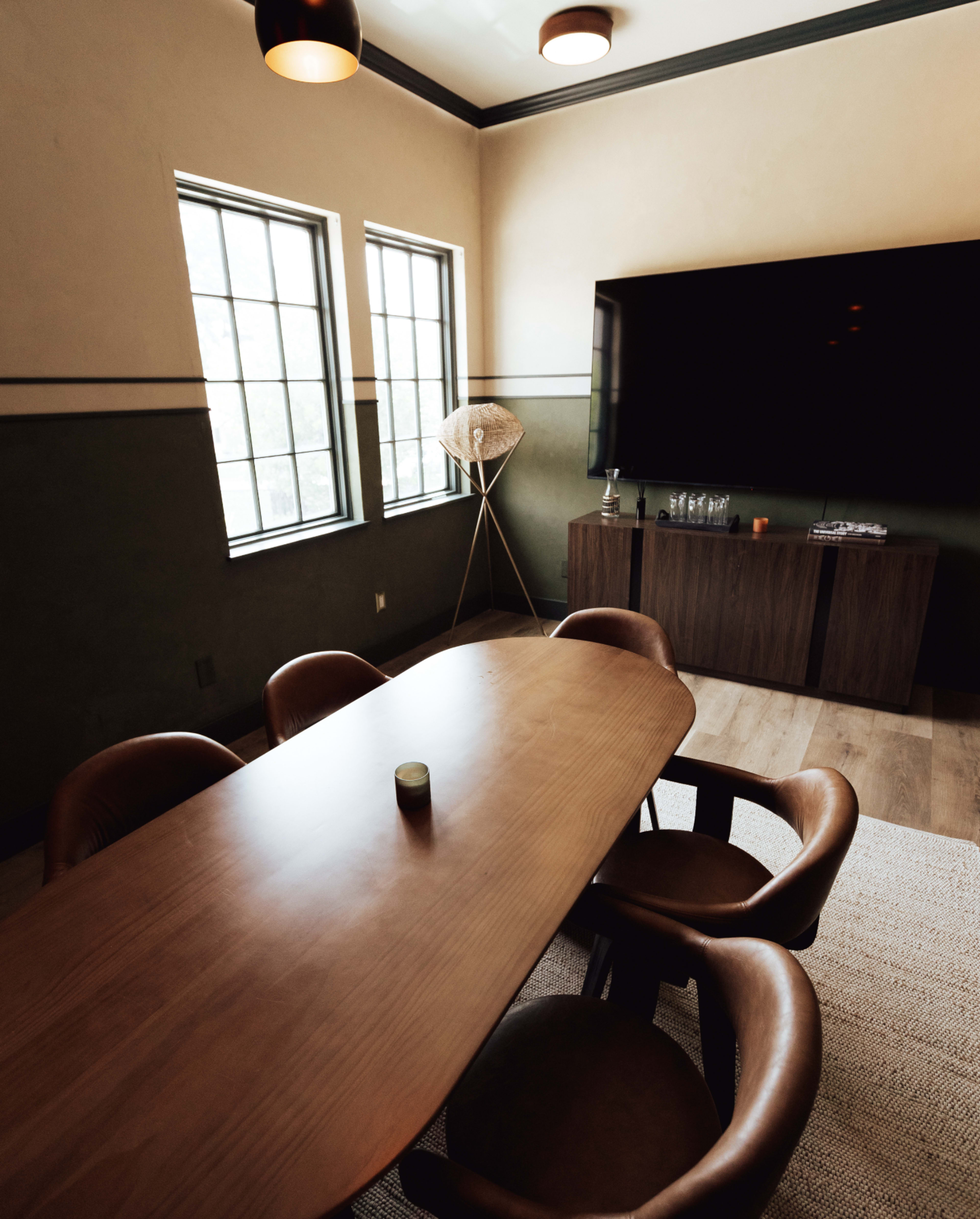 A rectangular wooden dining table with circular chairs is set in a room featuring two windows, a television, and a floor lamp.