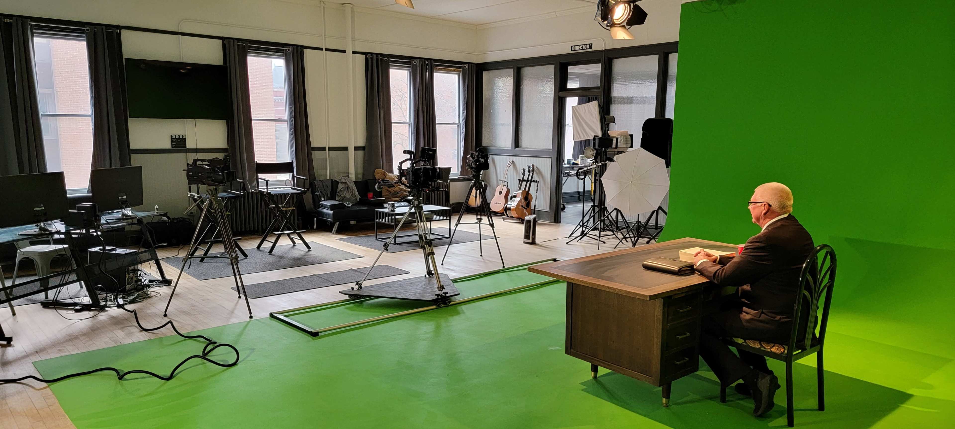 A man sits at a desk in front of a green screen in a brightly lit studio, surrounded by cameras and lighting equipment.