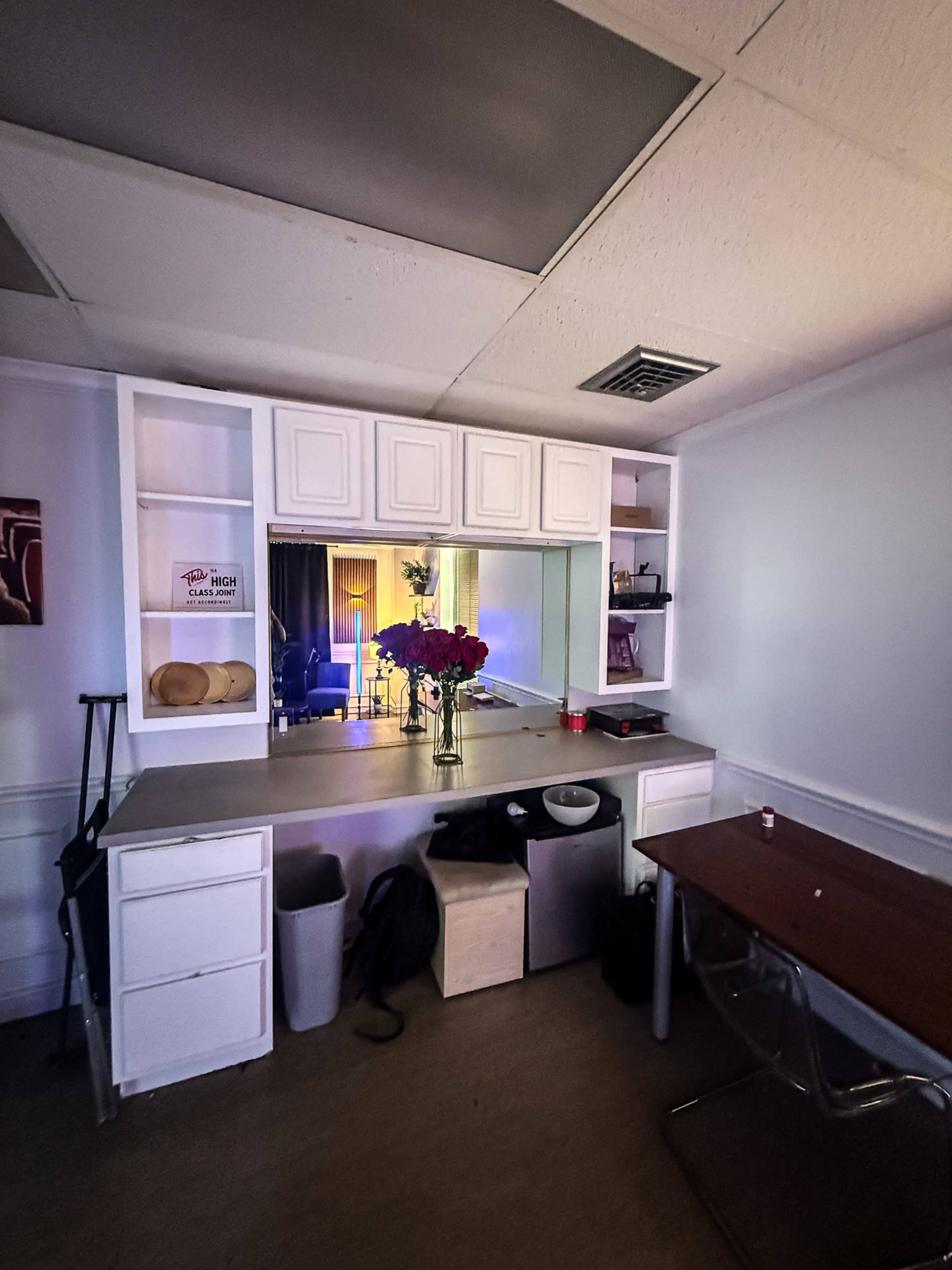 The image shows a small room with a counter area featuring shelves, a vase of flowers, and a chair beside a table.